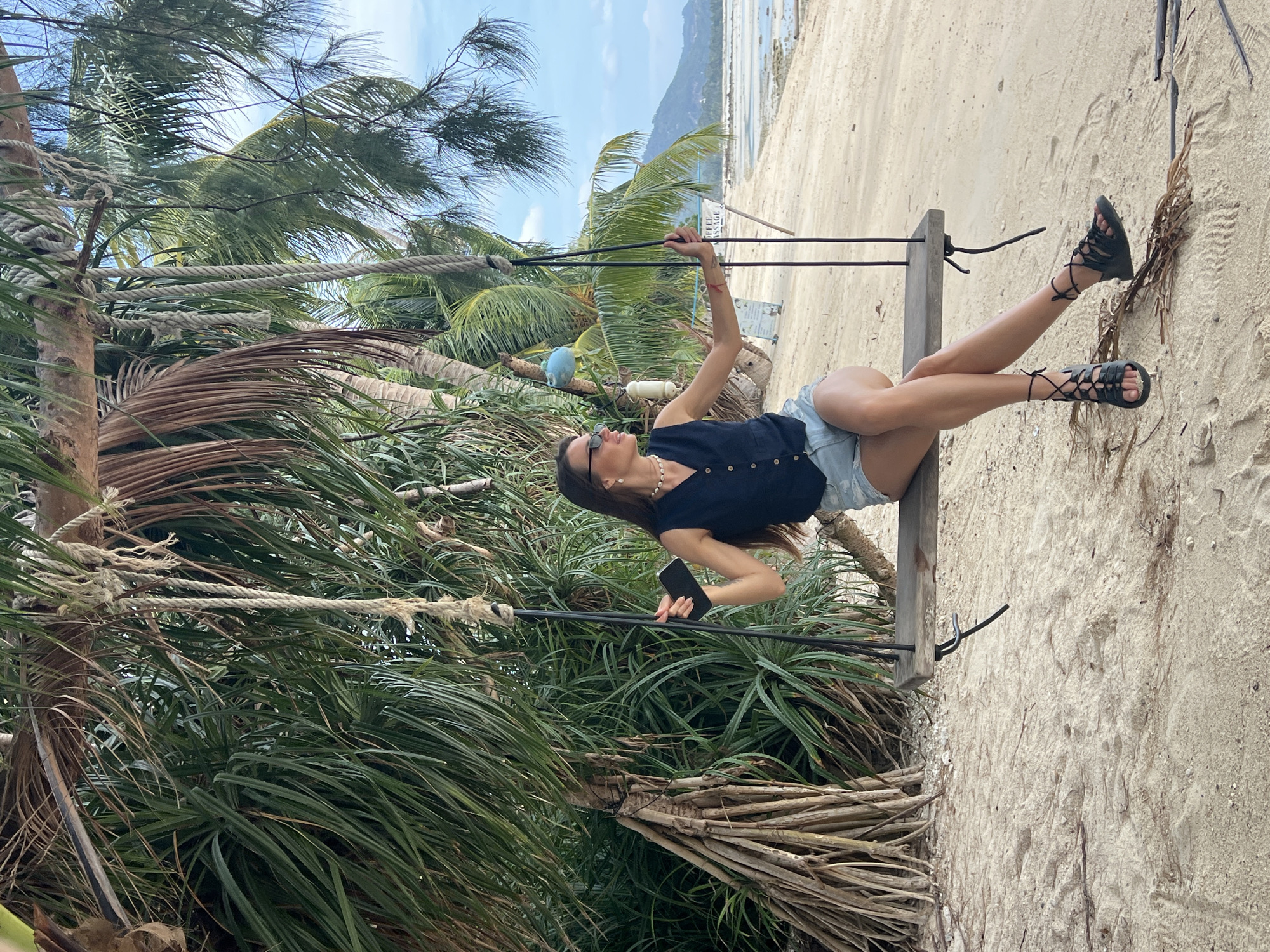 The image presents a serene tropical beach scene featuring a young woman posing on a rustic swing. The photograph is currently oriented vertically, appearing rotated 90 degrees clockwise from a natural upright view, with the woman lying across the frame.
The main subject is a woman, likely in her 20s or 30s, with long brown hair. She is dressed in a dark blue sleeveless top with buttons, light-colored denim shorts, and black lace-up sandals. Accessories include sunglasses and a light-colored beaded necklace. She is lying on her back on a wooden plank swing, which is suspended by thick natural fiber ropes. Her head points towards the left of the frame, and her legs are extended towards the right. Her right arm is raised, holding onto one of the swing's ropes, while her left hand holds a dark-colored smartphone. She appears relaxed, gazing upwards.
The setting is a lush beach environment. The foreground and midground are dense with tropical vegetation, including tall palm trees with visible fronds and trunks, as well as thick, spiky green foliage, some with dried brown fronds. The swing's ropes are visible, robustly tied around tree trunks. Beyond the vegetation, a sandy beach stretches into the background, showing footprints and natural undulations. In the far background, a calm, light blue sea meets the horizon, with distant green hills or mountains under a bright, partly cloudy sky. The lighting suggests a clear, sunny day, likely morning or early afternoon.
Notable details include a partially visible white sign with black text in the background, displaying "DEFE-" and "ASSAGE," indicating a possible spa or massage service. Two small objects, one blue and one white, are also visible attached to a tree branch near the woman's arm. The overall atmosphere evokes leisure and vacation in a tropical locale, consistent with Baan Thong Po, Thailand.
