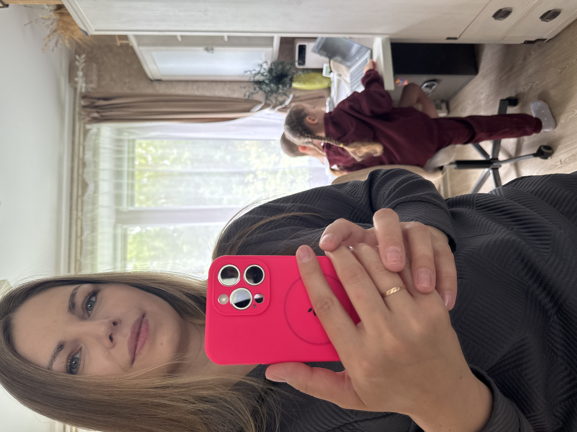 A woman is taking a selfie indoors, holding a pink smartphone with a visible camera lens. Two children, one with blonde braids, are sitting at a desk behind her, appearing to work on a laptop. The environment includes a window with natural light, a desk with computer equipment, and a drawer unit. The image suggests a domestic setting. The scene is in Раздолье, Russia.