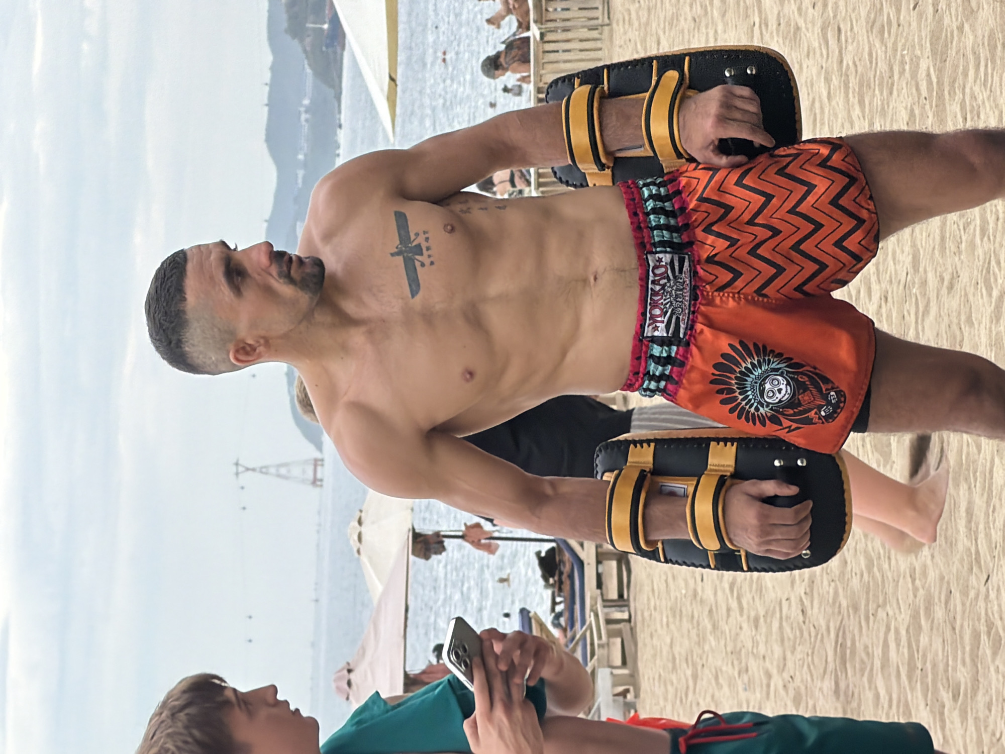 A man with a shaved head and a goatee, wearing orange Muay Thai shorts with a black chevron pattern and a tribal design, stands on a sandy beach in Nha Trang, Vietnam. He is shirtless, displaying a tattoo on his chest, and is wearing black and yellow Muay Thai pads on his forearms. Another person, partially visible on the left, is wearing a green shirt and holding a smartphone. In the background, beach umbrellas, lounge chairs, and the ocean are visible. The sky is overcast.