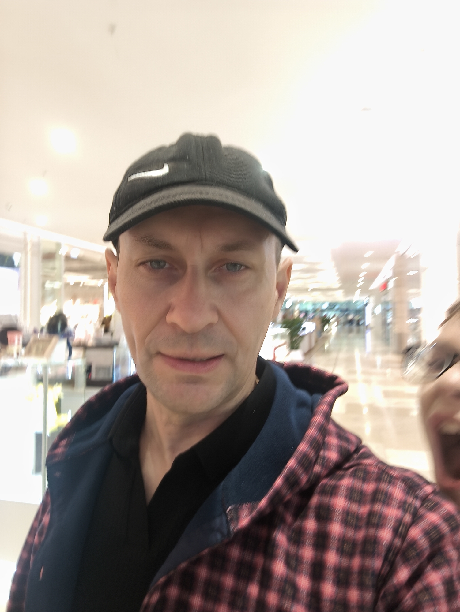 A man wearing a black baseball cap with a white Nike logo and a red and black plaid shirt over a black shirt stands in what appears to be an indoor shopping mall. The man is looking at the camera, and his mouth is slightly open. In the background, a blurred image of another person's face is visible, with their mouth open in a smile. The shopping mall appears to be brightly lit, with several storefronts visible in the distance. The overall image is slightly blurry, suggesting it was taken in motion or with a low-resolution camera. The location is not specified as Vladivostok, Russia.