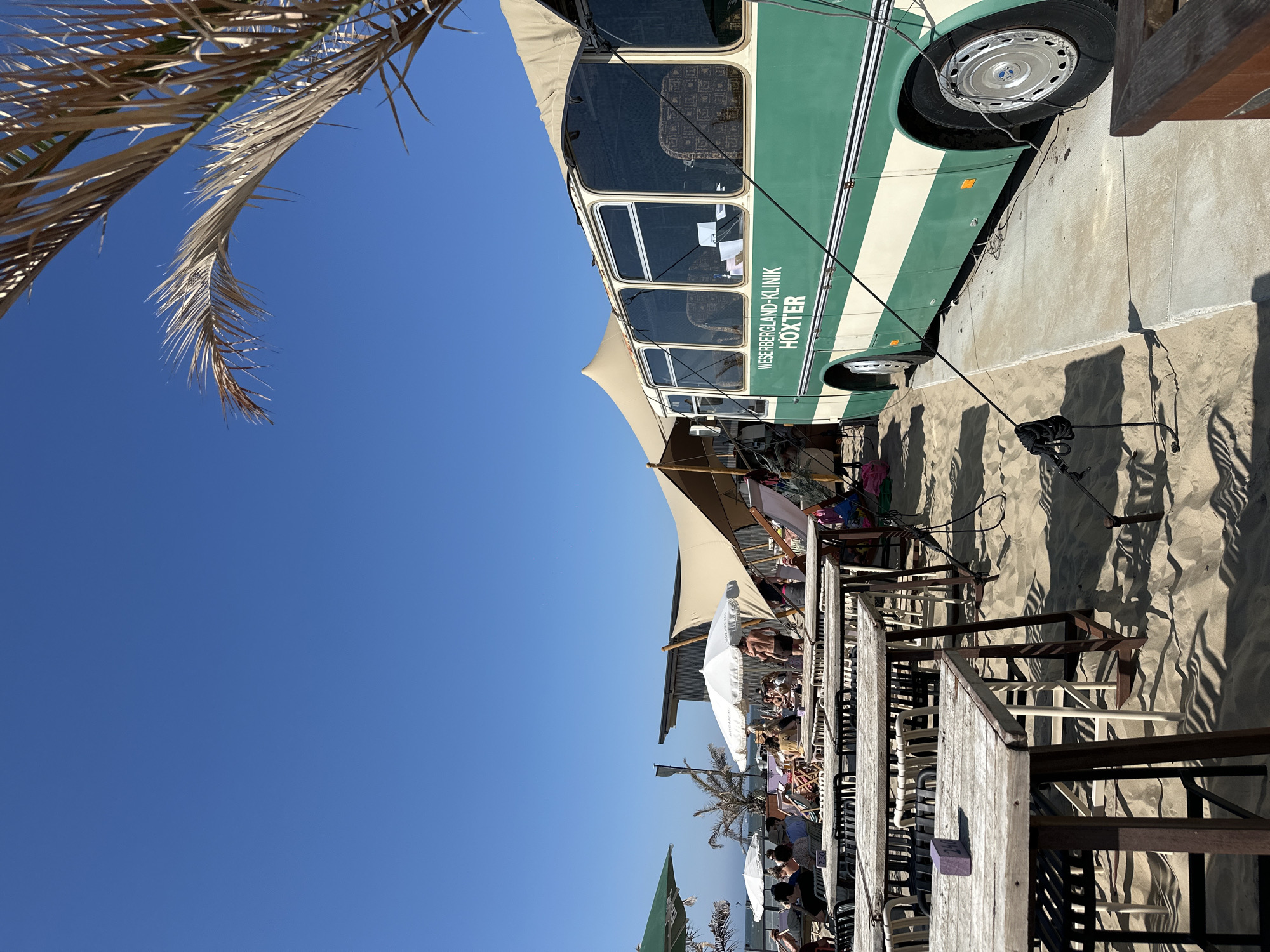 This image captures a sunny day at a beachside establishment in Zandvoort, Netherlands. In the foreground, a row of empty wooden tables and chairs on a sandy beach are bathed in sunlight, casting long shadows. To the right, a vintage green and cream bus, identified with the text "WESERBERGLAND-KLINIK HÖXTER," is parked on a concrete ramp. The bus appears to be part of the establishment, possibly serving as a bar or dining area.
In the background, a large, beige, tent-like structure offers shade over a seating area with white umbrellas and numerous people. A few palm trees, one dry and sparse, add to the beachy atmosphere. The clear blue sky suggests fair weather. The scene depicts a relaxed outdoor dining or lounge area by the sea, with a unique bus as a central feature.