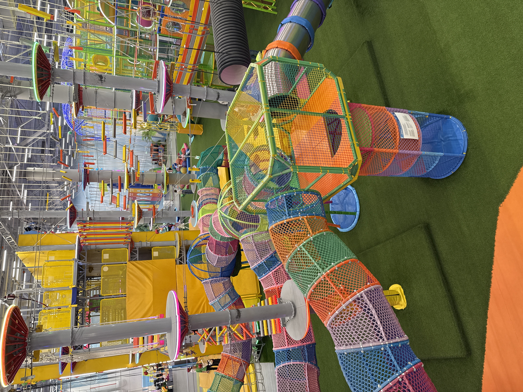 The image depicts a colorful indoor playground structure located in Khimki, Russia. The playground features a network of tunnels, slides, and climbing structures made of netting and metal. The tunnels are cylindrical in shape and are connected to each other by various platforms and passageways. The main colors are green, yellow, red, orange, pink and blue. The entire structure is designed to provide a fun and engaging environment for children. The playground is set against a backdrop of other playground equipment and elements, creating a visually stimulating and playful atmosphere.