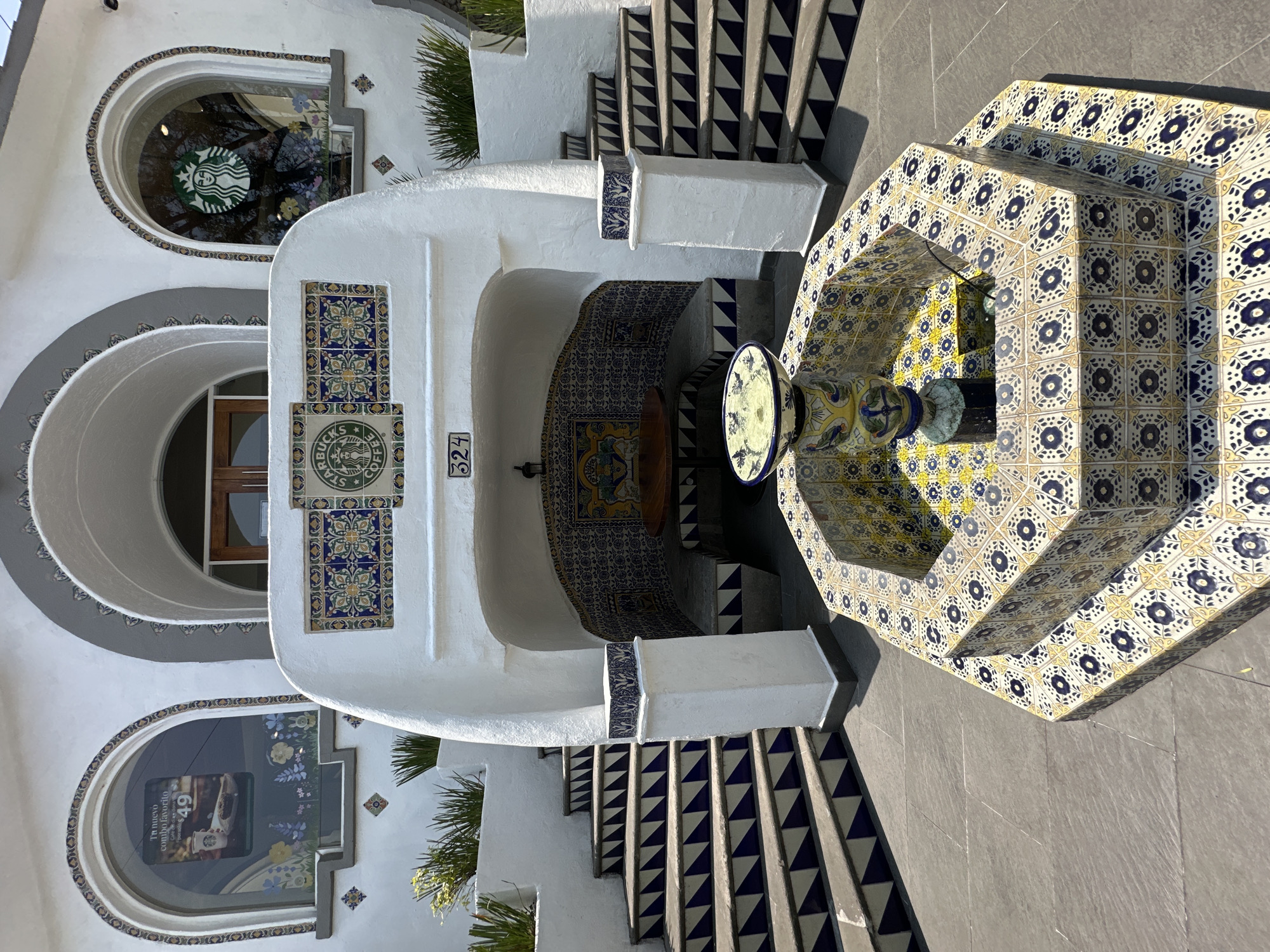 This media file captures a vibrant and ornate architectural scene, likely an entrance or courtyard, characteristic of traditional Mexican design. The scene is brightly lit by natural sunlight, casting sharp shadows and indicating a clear day. No people are visible.
The primary subjects are a white stucco building, a multi-tiered fountain, and a decorative staircase, all extensively adorned with colorful Talavera-style tiles.
On the left, the white building features distinct arched windows. One upper window clearly displays a green and white Starbucks logo. Below it, another arched window shows a promotional poster with text, "Tu nuevo combo favorito" (Your new favorite combo) and the number "49". Various square and rectangular tile mosaics, predominantly in blue, yellow, and green floral patterns, decorate the building's facade.
In the center, a prominent white archway or recessed entrance is visible, featuring a tiled bench or wall within. The address number "324" is displayed vertically on the wall to the right of this archway. A smaller, circular tile with the Starbucks siren logo and "STARBUCKS COFFEE" text is integrated into the tiling near the arch.
To the right, a tiered octagonal fountain stands out, completely covered in intricate blue and yellow geometric and floral patterned tiles. Water is visible in its basin. Adjacent to the fountain, a staircase ascends, with its risers decorated in a repetitive blue and white triangular pattern, complementing the overall aesthetic.
The setting evokes a welcoming and culturally rich atmosphere, with the pervasive tile work being a key visual element and a strong location cue for Mexico City, Mexico.