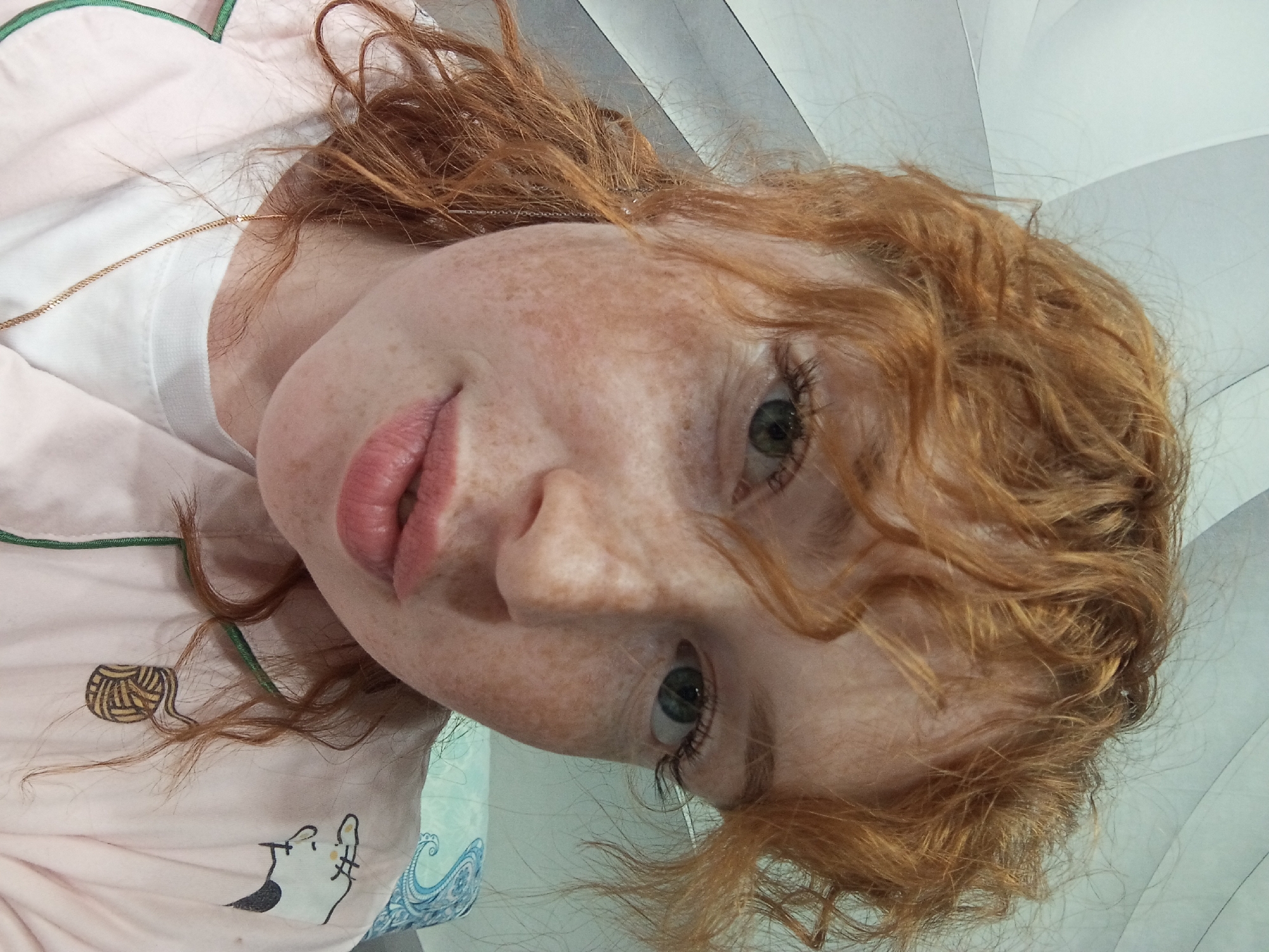 A close-up, slightly angled portrait shows a fair-skinned person with red curly hair and freckles. The person is wearing a light pink shirt with a white collar and a green trim. The shirt has cartoon-like designs on it, including a cat and a ball of yarn. A delicate gold chain necklace is visible. The background is a gradient of light gray and white. The location cannot be determined from the given image.