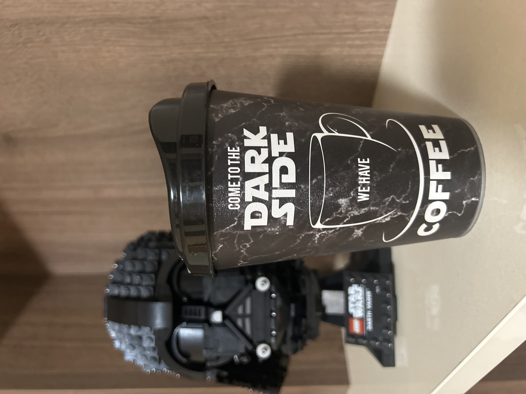 The image, taken in Veliko Tarnovo, Bulgaria, showcases a black and white coffee cup resting on a Lego Darth Vader helmet, set against an indoor backdrop. The photograph is oriented sideways, with the items appearing horizontally.
The prominent subject is a dark-colored reusable coffee cup or tumbler. It features a black background with a subtle white marbling or textured pattern. White text on the cup boldly declares: "COME TO THE DARK SIDE WE HAVE COFFEE." Between "SIDE" and "WE HAVE," there is a simple, stylized white outline of a coffee mug. The cup is fitted with a black lid.
Beneath the coffee cup is a meticulously constructed Lego model, specifically the iconic black helmet of Darth Vader from Star Wars. The intricate detail of the Lego bricks forming the helmet is visible. A small display plaque at the base of the Lego model is partially visible, showing the text "LEGO STAR WARS DARTH VADER."
The background consists of a light brown wooden surface extending across the left side of the frame, suggesting a shelf or tabletop. To the right, a lighter, possibly off-white, vertical surface is visible, implying a wall or the back of a cabinet. The scene is static, with no discernible activity occurring. The lighting appears to be soft and ambient, consistent with an indoor setting.