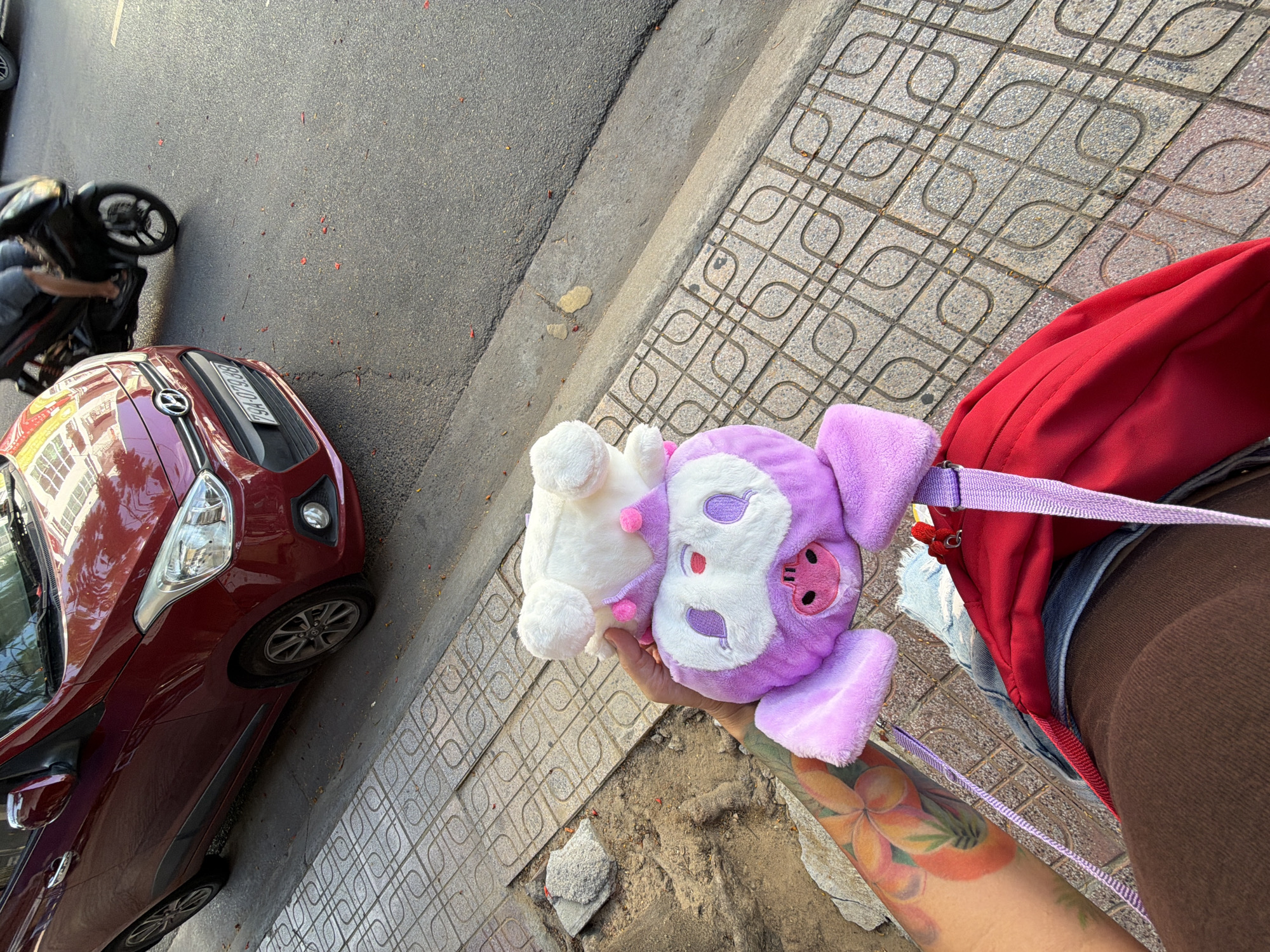 A street scene in Nha Trang, Vietnam, shows a red car parked on the right side of the road, with a motorcycle partially visible in the background. In the foreground, a person is holding two stuffed toys, a white one on top of a purple one, while wearing a red backpack and brown pants. The person has a colorful tattoo on their left arm. The sidewalk is paved with square tiles featuring a patterned design, and the street is asphalt. Red flower petals are scattered on the road and sidewalk.