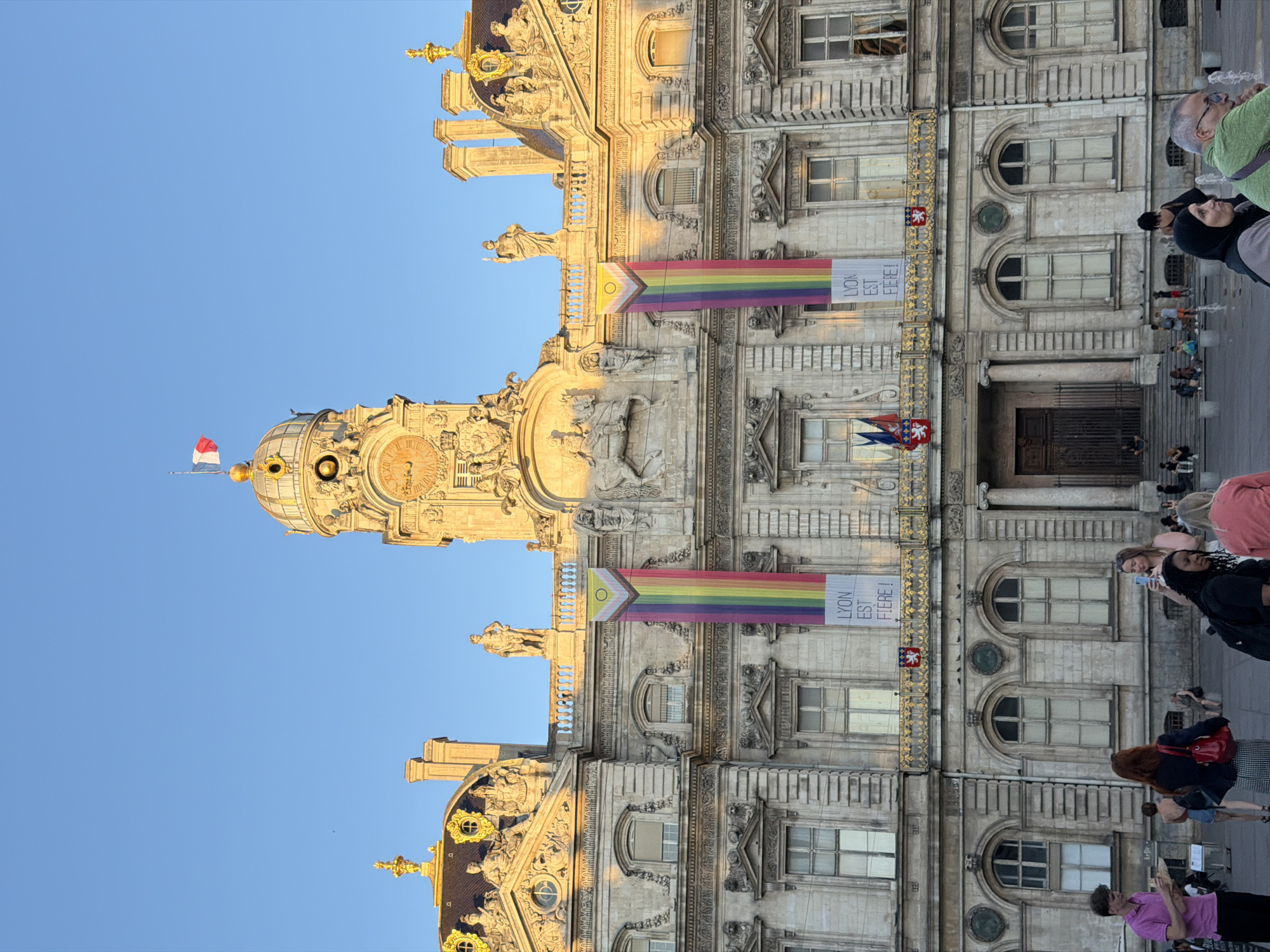The image shows the Hôtel de Ville (City Hall) of Lyon, France, a grand stone building with classical architectural elements. A large clock tower is prominently featured at the center of the facade, topped with a French flag. Two large, vertical banners displaying the LGBTQ+ pride flag hang from the building's upper level, with text on the right banner reading "LYON EST FIÈRE!" (Lyon is Proud!). The building is bathed in the warm light of what appears to be a sunset. In the foreground, a number of people are visible, some standing, others sitting, suggesting a public gathering or a casual scene in a square in front of the city hall. The sky is clear and blue.