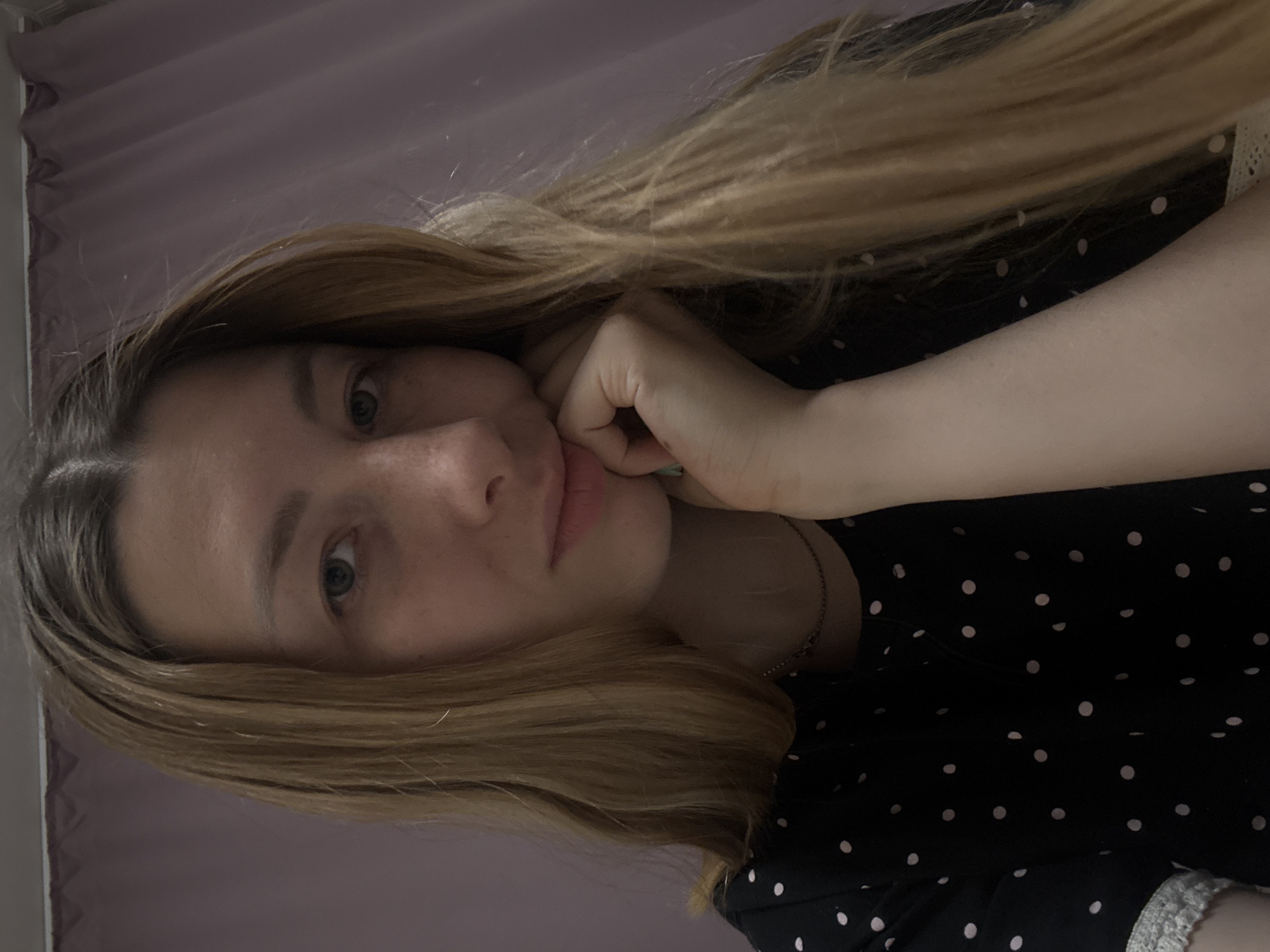 A woman with light-brown hair is resting her chin on her hand while facing the camera. She is wearing a black polka-dot dress. The background is a light-purple wall. There is no indication of a specific location in Moscow, Russia.
