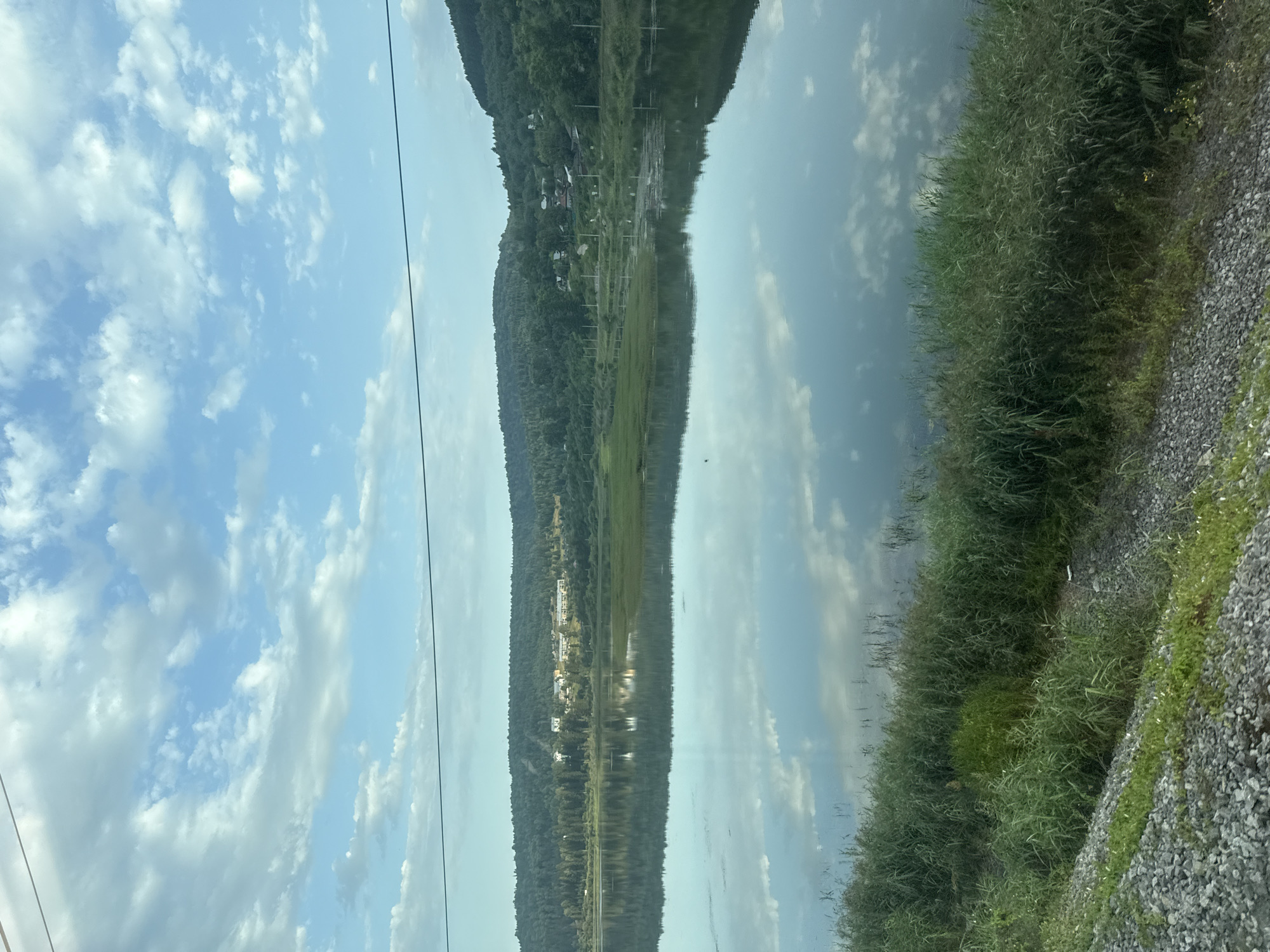 The image depicts a tranquil landscape scene with a body of water reflecting the sky and surrounding hills. The water's surface is calm, with the clear blue sky and scattered white clouds beautifully mirrored. Along the far bank, a lush green hillside is covered in dense forestation, with a few buildings visible nestled amongst the trees, suggesting a small settlement or village. In the foreground, tall green grasses and reeds grow along the water's edge, with a rocky embankment to the left of the frame. Power lines are visible crisscrossing the sky, indicating the presence of infrastructure. The overall impression is one of natural beauty and serenity, possibly captured from a moving vehicle, given the slight blur in the foreground and the perspective of the shot. The location is identified as Minyar, Russia.