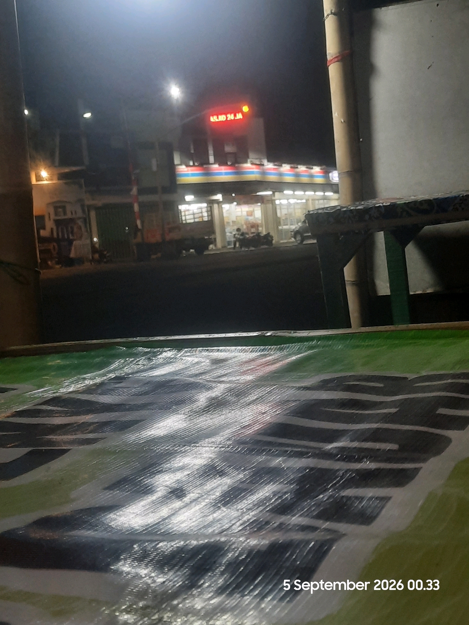 The image captures a nighttime scene on a street in Sukokerto, Indonesia, in front of a brightly lit convenience store. The store's facade features prominent red neon lettering that reads "ASLI 24 JA" and horizontal stripes of red, white, and blue above rows of windows. Several vehicles, including motorcycles and cars, are parked in front of the store, and some individuals are visible inside through the windows. The street is dark, with illumination primarily coming from the store's lights and streetlights. In the foreground, a large green and black tarp is spread out, partially obscuring the view of the street. The tarp has a glossy, wrinkled surface that reflects the ambient light. The overall impression is one of a typical urban street scene at night, with the convenience store serving as a central point of activity. The date and time stamp "5 September 2026 00.33" is visible in the bottom right corner of the image.