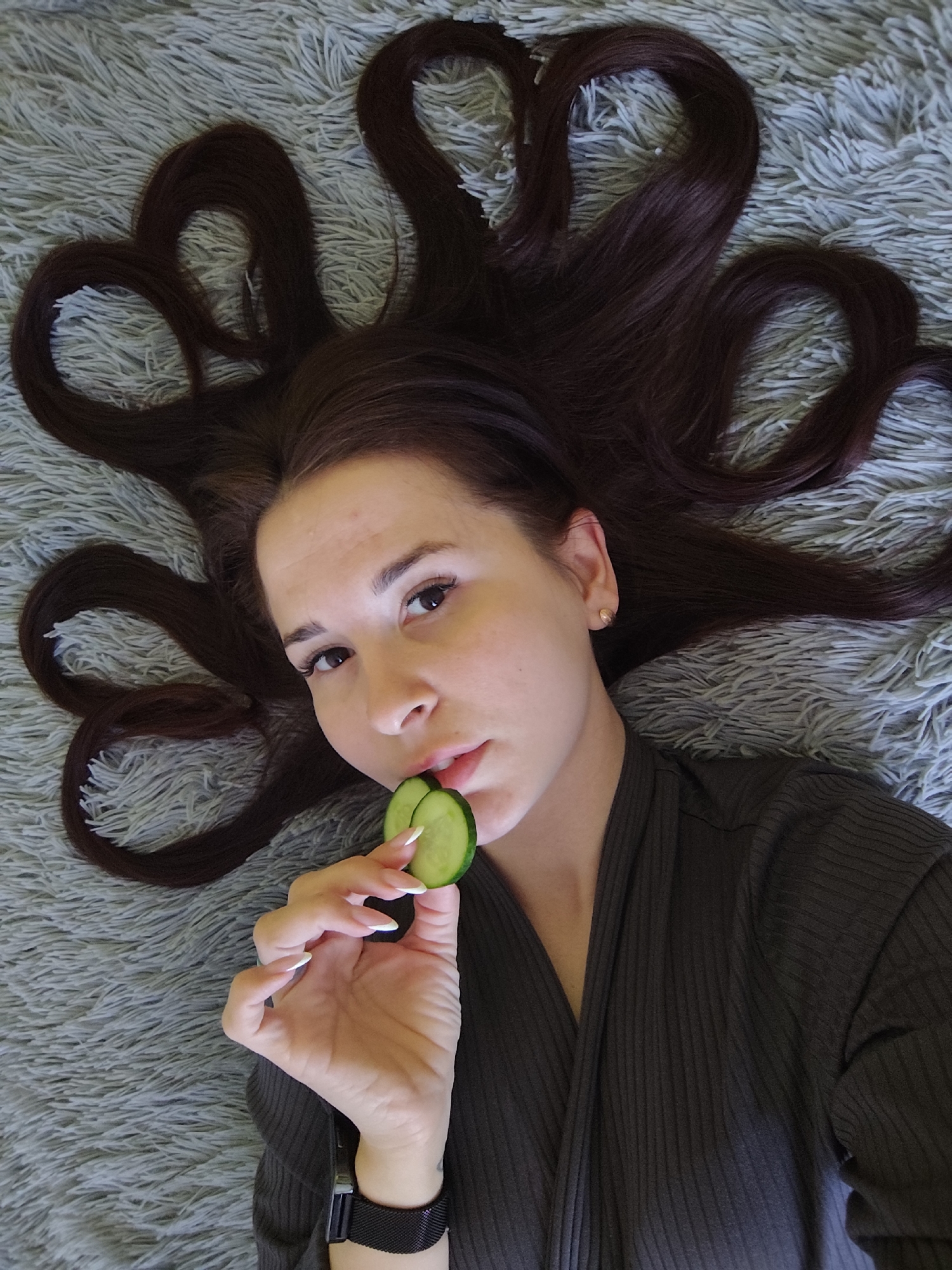 A young woman lies on a fluffy grey rug with her dark brown hair spread out around her head in the shape of hearts. She is holding a slice of cucumber near her lips, appearing as if she is about to eat it. The woman wears a dark-colored, ribbed top or robe. Her fingernails are long and painted with a white tip. She has a watch with a black band on her left wrist. The location of Фоки, Russia, is not discernible from the visual information provided in the image.