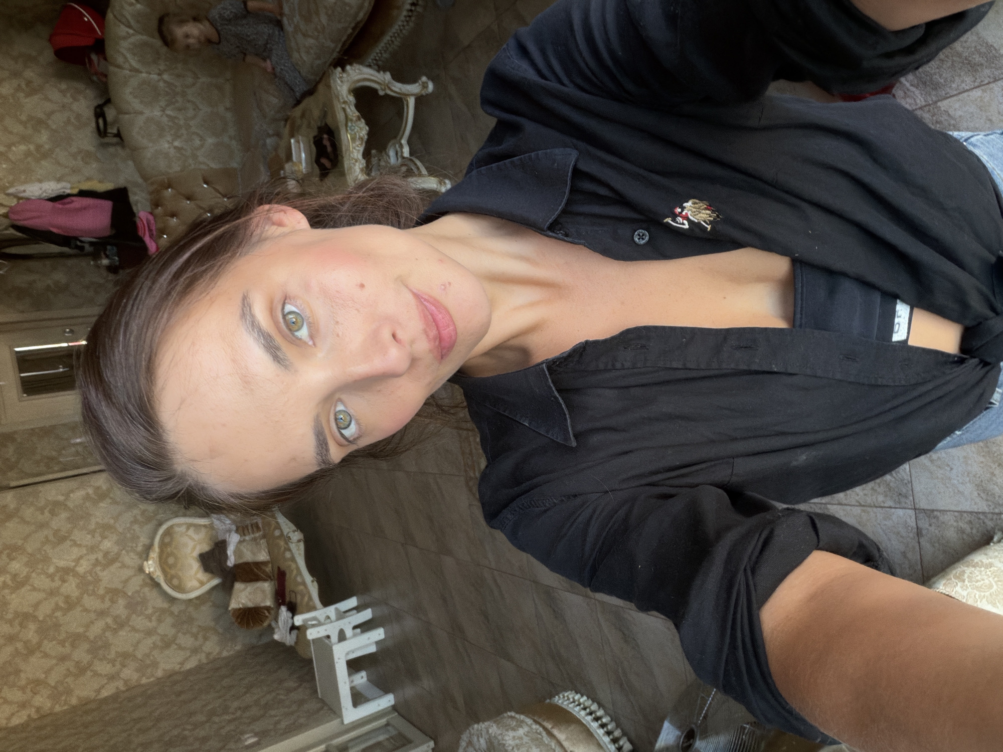 A woman with light brown hair and green eyes is taking a selfie in a room with ornate furniture and patterned wallpaper. She is wearing a black unbuttoned shirt over a black bralette and blue jeans. A small, embroidered logo is visible on her shirt. In the background, a child in a grey and white patterned outfit is sitting on a couch. The floor appears to be tiled. The lighting suggests it is daytime. The overall impression is of a domestic setting, likely a living room. The location is Ufa, Russia.
