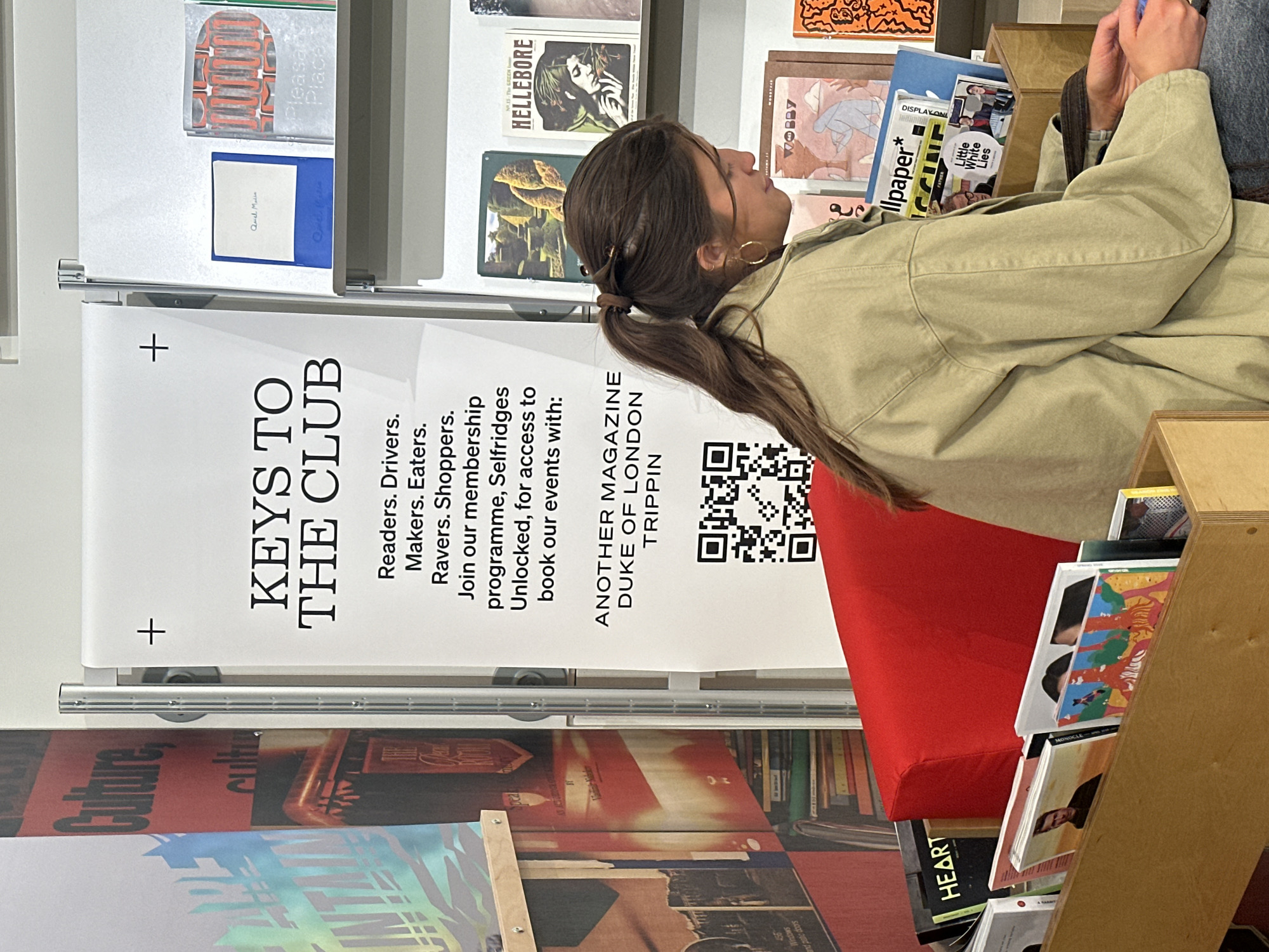 The image shows the interior of a shop, likely a newsstand or magazine store in London, United Kingdom. A woman with long brown hair tied back in a ponytail, wearing a beige jacket and gold hoop earrings, is sitting on a red cushion chair, looking at magazines. A banner with the words "KEYS TO THE CLUB" is prominently displayed, advertising a membership programme with Selfridges and Another Magazine at the Duke of London, Trippin. The banner features a QR code. Various magazines are visible on the shelves, including "Wallpaper*", "Little White Lies," "Hellebore," and "Heart." The "Culture" banner and some books and objects are visible on the other racks above the KEYS TO THE CLUB ad.