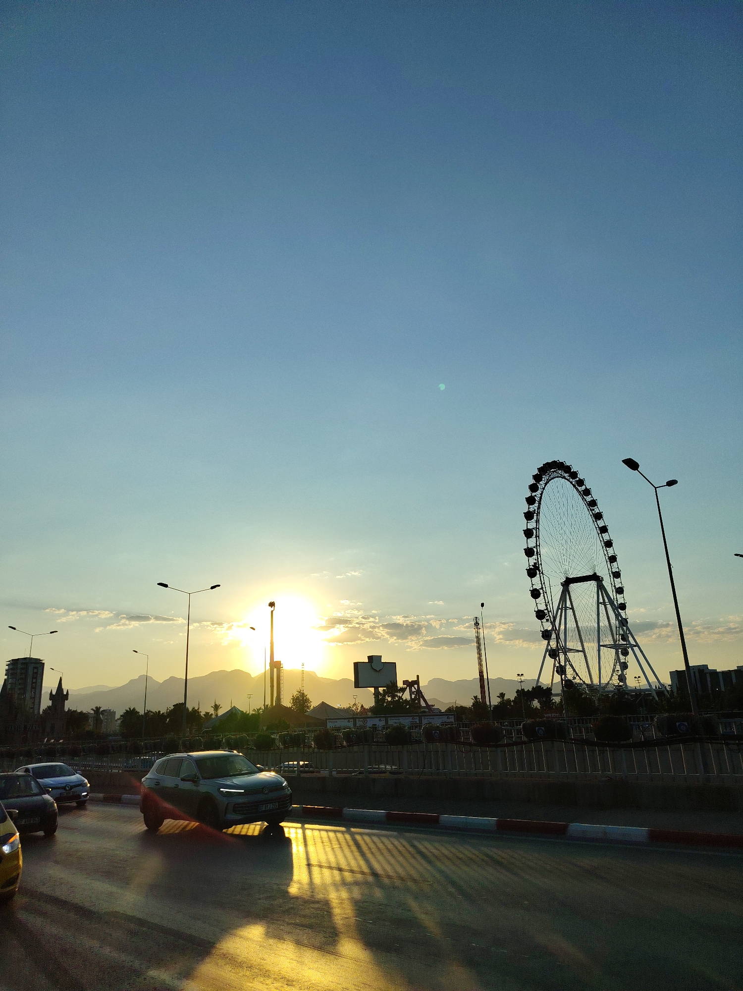 The image depicts a sunset scene in Konyaaltı, Turkey, with a prominent Ferris wheel silhouetted against the sky. The sun is low on the horizon, casting a warm golden glow across the scene and creating long shadows. Several cars are visible on a road in the foreground, with one silver SUV prominently featured. In the background, mountains form a distant backdrop, and buildings and streetlights can be seen. The sky is a gradient of blue and yellow, with scattered clouds.
