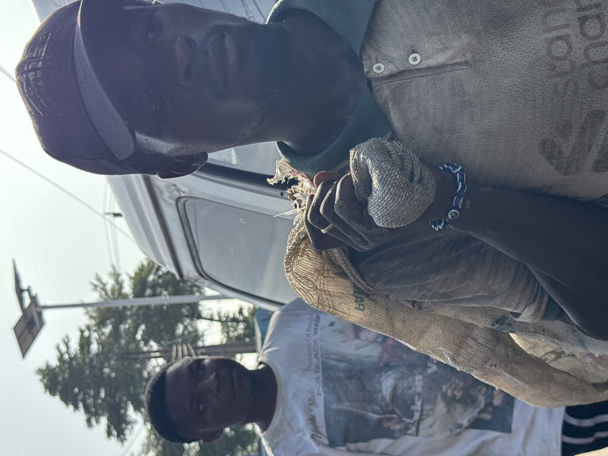 In Ado, Nigeria, the image shows two Black men. The man closest to the camera is wearing a dark cap and a light-colored button-down shirt, with a beaded bracelet on his wrist. He is holding a frayed, woven material. A second man stands behind him, wearing a white t-shirt with an image printed on it. A white van is visible in the background. The scene appears to be outdoors during the day, with trees and a traffic signal visible in the background.