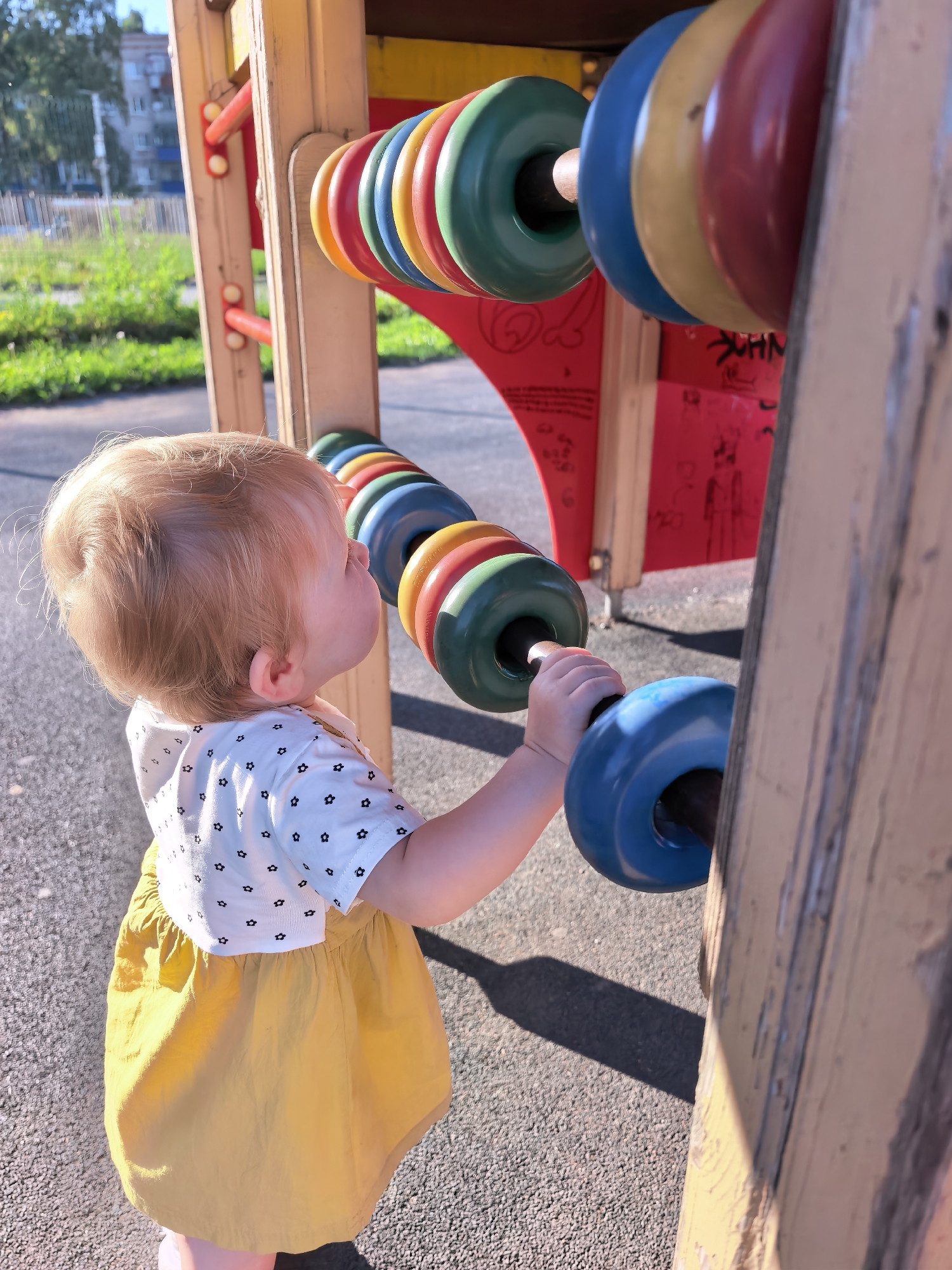 A young child with blonde hair, wearing a white shirt with black star patterns and a yellow skirt, plays with a colorful abacus on a playground. The abacus has rows of large, brightly colored beads, including blue, green, yellow, and red. The child's left hand is reaching out to touch a blue bead. The playground equipment is made of wood and painted in yellow and red. In the background, greenery and a building are visible, suggesting an outdoor urban setting. The ground is paved with a textured surface. The lighting suggests it is a sunny day.