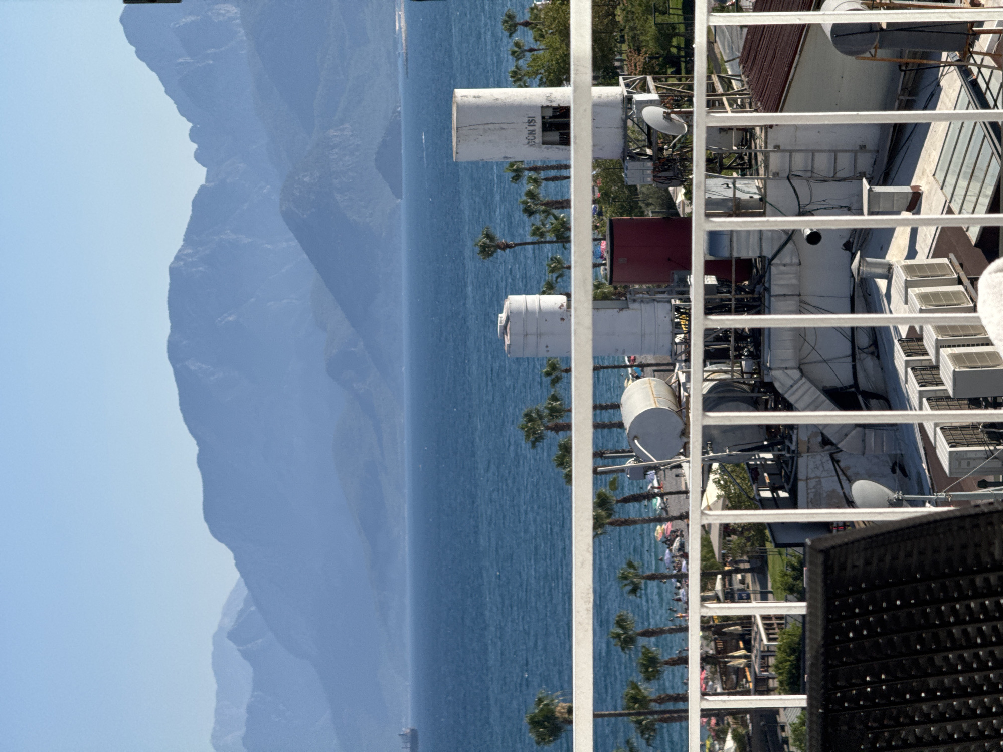 This is a vertically oriented, eye-level, medium shot of a coastal scene. In the foreground, a white railing runs horizontally across the frame, obscuring part of the view. Beyond the railing, to the left, are layers of blue water and hazy blue mountains. The water is calm, with small ripples visible. Palm trees are scattered along the shoreline, and further in the distance, there appears to be a large ship.
To the right of the water and mountains, and behind the railing, are various rooftop structures. These include what appear to be air conditioning units, satellite dishes, and cylindrical tanks. One of these tanks has text on it that reads "GÜN İŞİ." There are also some red and white structures, possibly part of buildings. Below the rooftop equipment, a section of beach is visible with numerous people, some lying on towels and umbrellas, suggesting a sunny day at the beach. The overall impression is a view from a balcony or rooftop overlooking a bustling beach scene with a mountainous backdrop, likely in a warm climate. The time of day appears to be daytime, with clear skies suggested by the bright lighting and the haze over the distant mountains.