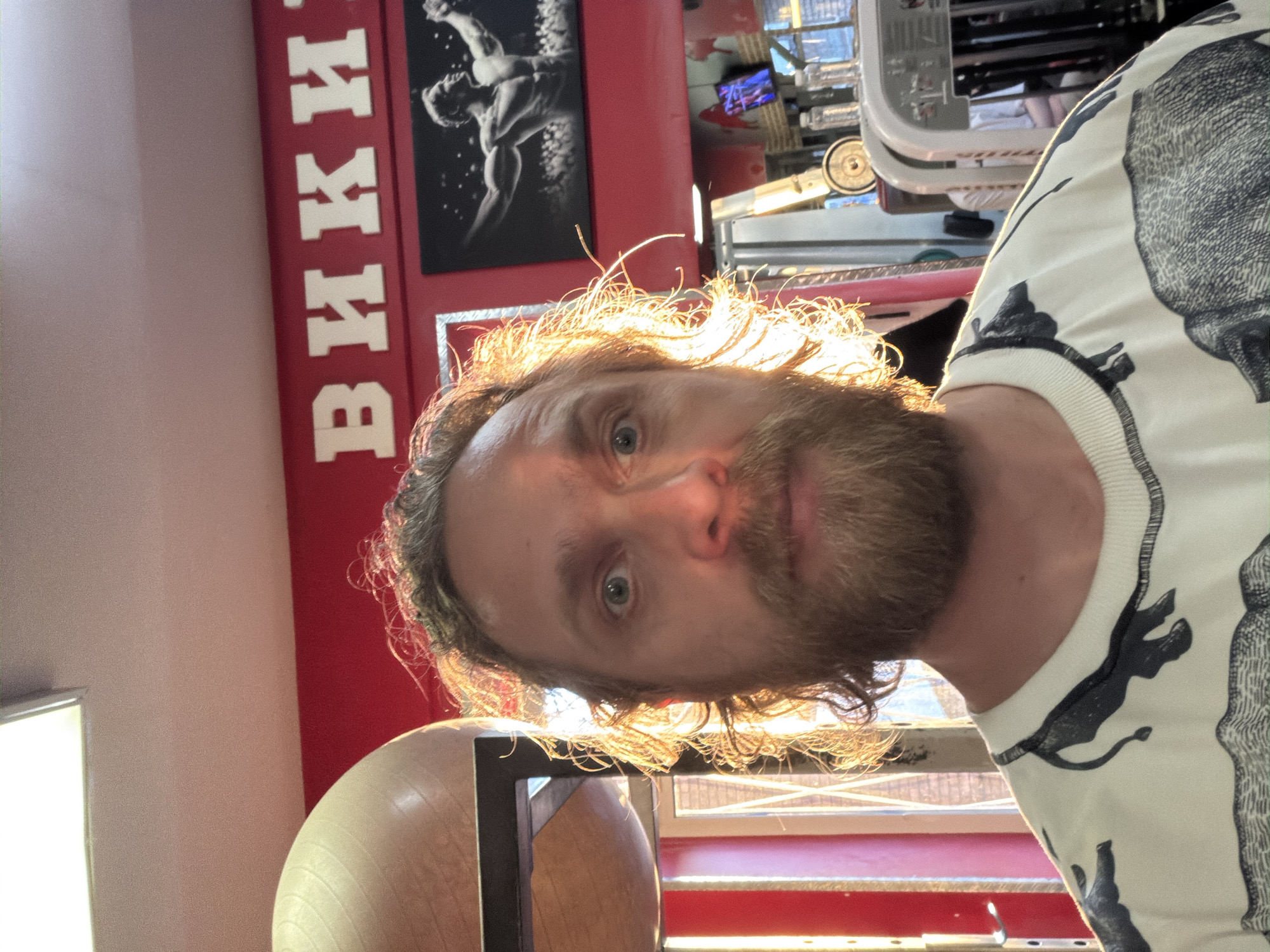 The image displays a man with a beard, mustache, and medium-length wavy hair, looking directly at the camera with a neutral expression. His hair is backlit by strong, golden light, creating a halo effect, suggesting late afternoon or early morning sunlight. He is wearing a white sweatshirt with black stylized patterns resembling panthers or jaguars.
The setting is an indoor gym or fitness center, located in Saint Petersburg, Russia. To the man's left, a vibrant red wall features large white Cyrillic letters spelling "ВИКИ" (VIKI/WIKI). Above this text, a framed black-and-white picture of a muscular male figure in an athletic pose is mounted on the wall. Partially visible in the lower foreground is a light-colored exercise ball. Various metallic structures and gym equipment can be seen blurred in the background. The man appears to be taking a selfie or posing for a photograph.
