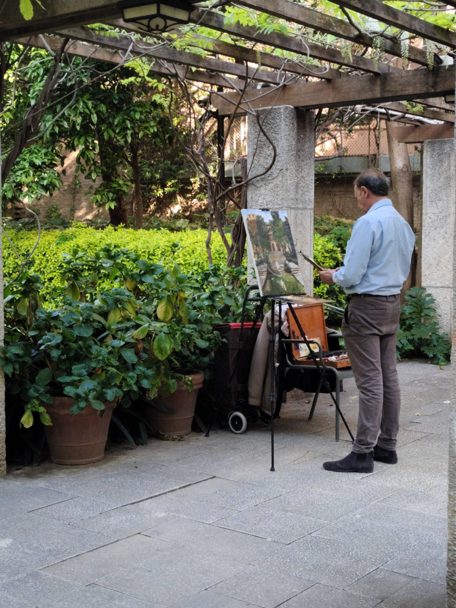 An artist stands painting outdoors in a garden or courtyard, possibly in Barcelona, Spain. He wears a light blue long-sleeved shirt, brown pants, and black shoes. He faces his easel, holding a paintbrush in his right hand, seemingly in the process of painting. The canvas on the easel displays an image of a European landscape with architecture, water, and greenery. His painting equipment, including a palette and possible paint supplies, are arranged on a chair beside the easel. The scene is situated under a pergola structure overgrown with vines and greenery. Several potted plants are in the foreground. The ground is paved with large, flat stones. A wall overgrown with foliage serves as a backdrop, hinting at an urban setting.