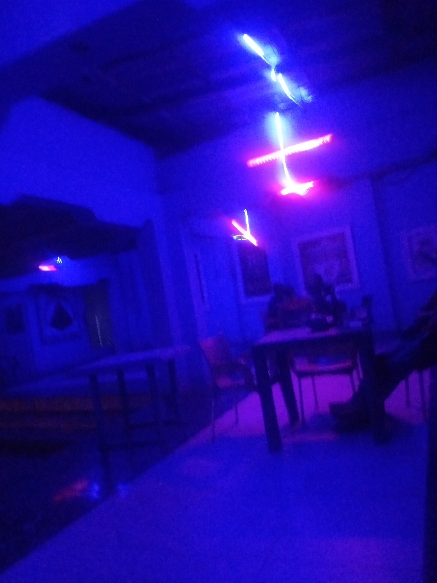 The image depicts an interior scene, possibly a lounge or entertainment venue, bathed in a vibrant blue and purple neon light. In the background, illuminated by the neon lights, are what appear to be decorative elements or installations hanging from the ceiling, forming abstract shapes and lines of light. A table and chairs are visible in the mid-ground, with indistinct figures seated or standing around it. The flooring is a solid color, likely also reflecting the neon illumination. The overall ambiance is one of low light and artificial illumination, suggesting a nightlife or recreational setting. The location of Sabon Gari Nassarawa, Nigeria, is not directly discernible from the visual elements of the image itself but is provided as contextual information.