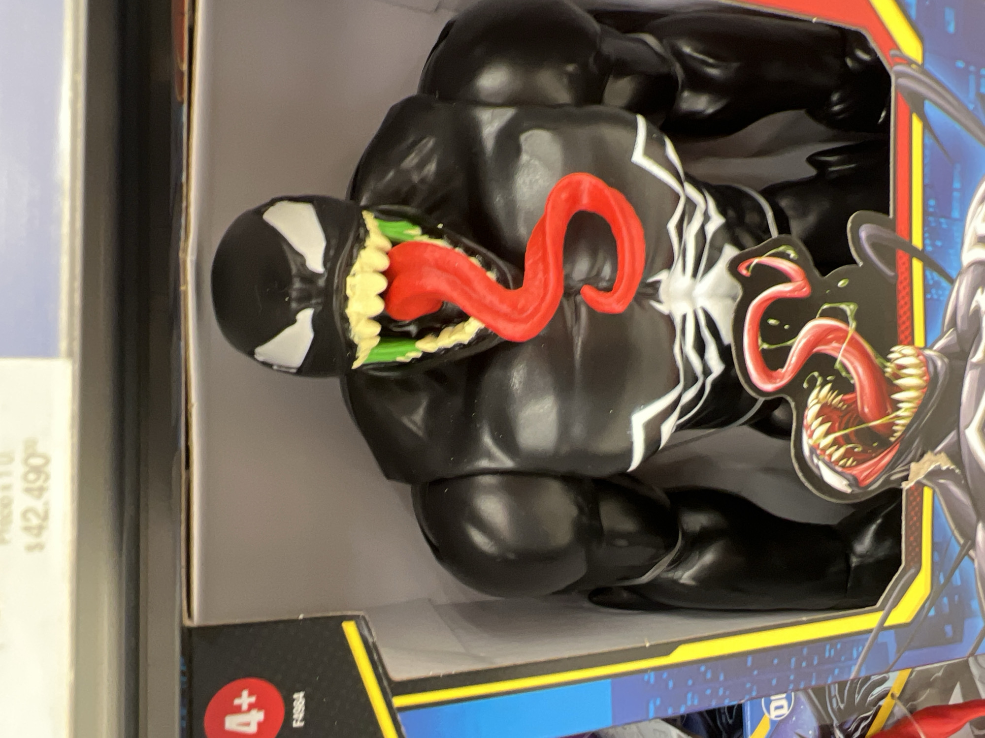 A Venom action figure is in a box. The toy is predominantly black with white markings and a long, red tongue. It is lying on its back in the box. The box is decorated with blue and yellow markings. A sticker on the bottom of the box reads "4+". The price sticker $42,490 is only partially visible. The city of Buenos Aires, Argentina, is not identifiable in the image.