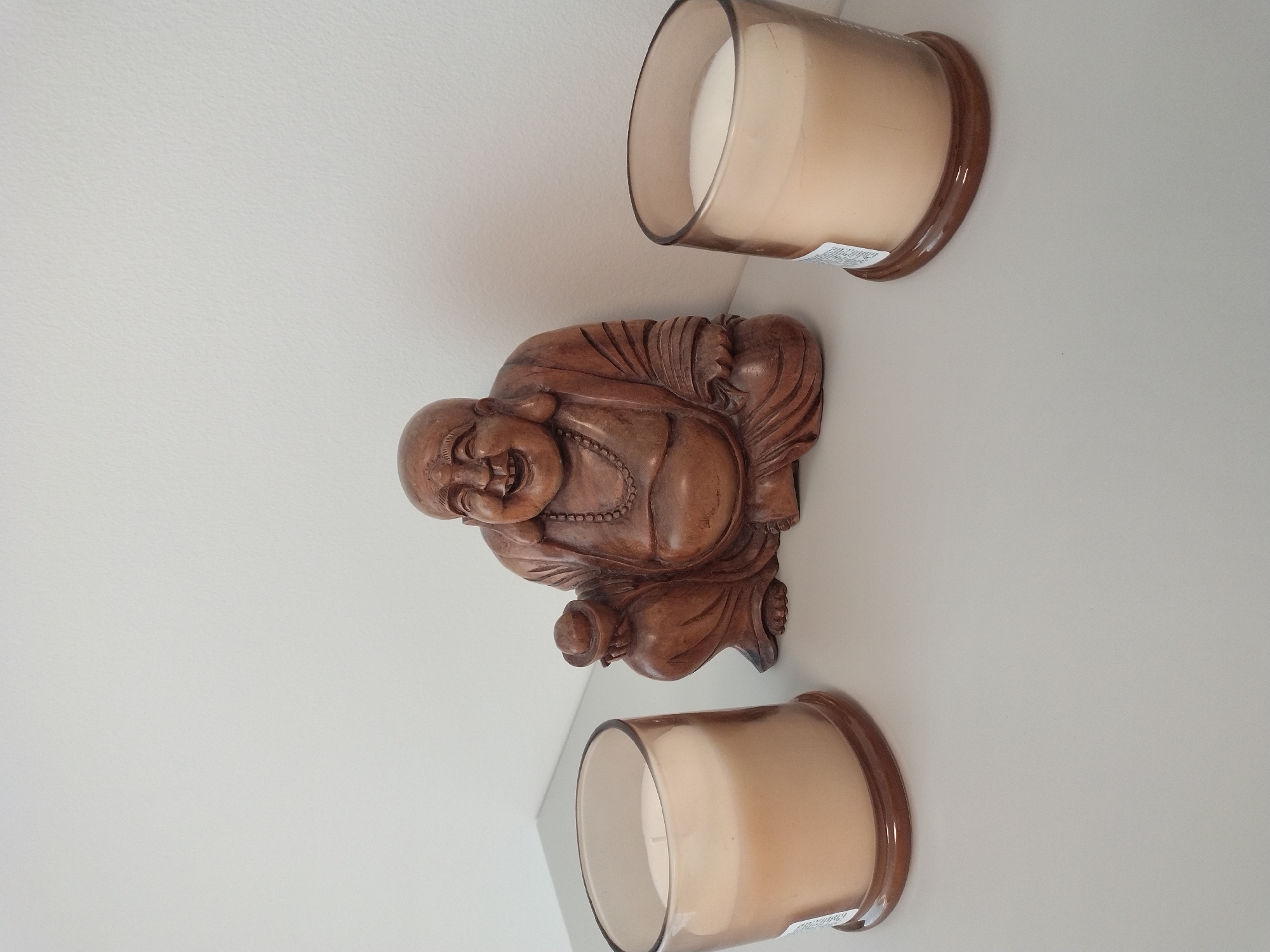 The image shows a wooden statue of a laughing Buddha figure situated between two lit candles in glass containers. The Buddha statue is centrally located, with a candle on either side. The candles appear to be a pale orange or cream color and are in clear glass jars with brown bases. The background is a plain, light-colored wall. The objects are placed on a flat, light gray surface. The scene is indoors and appears to be on a shelf or table. The location of this scene in Písek, Czechia, is not discernible from the image itself.