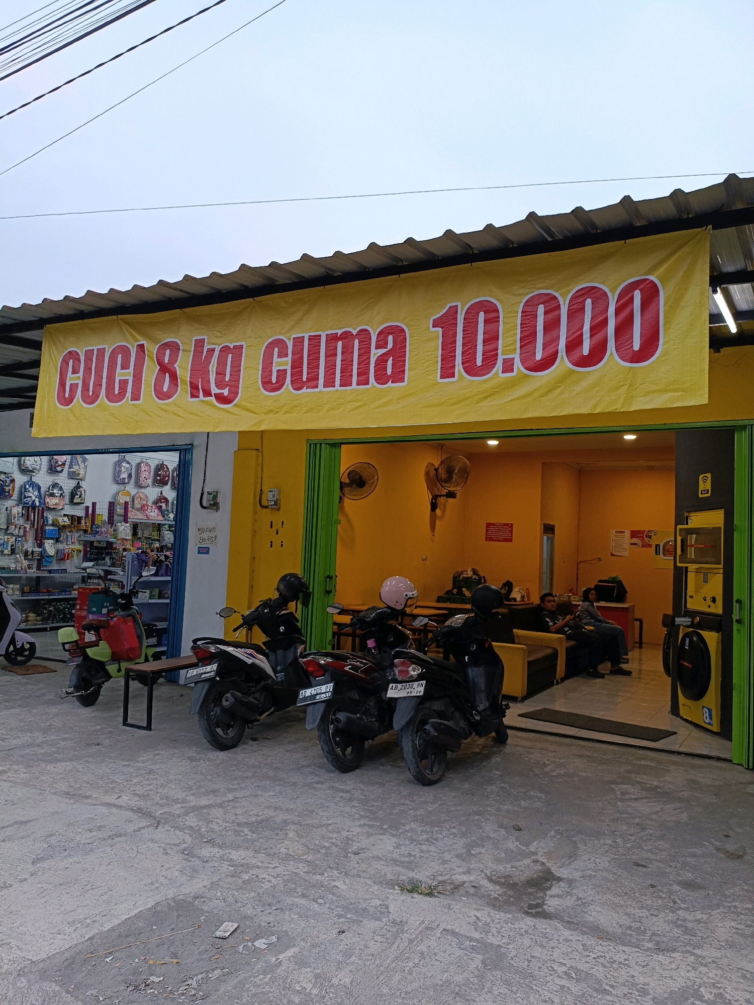 A brightly lit, open-air laundry service in Maguwoharjo, Indonesia, is prominently displayed with a large yellow banner proclaiming "CUCI 8 KG CUMA 10.000" in bold red lettering. The banner stretches across the top of the establishment, advertising a laundry service for 8 kilograms costing only 10,000 Indonesian Rupiah. In front of the service area, two motorcycles are parked on a concrete ground. The left motorcycle is a light green scooter, while the right motorcycle is a black motorbike.
The interior of the laundry service features yellow walls and is equipped with what appear to be washing machines and dryers, one of which is yellow and labeled with the number "8". Several people are seated on a yellow sofa inside, appearing relaxed, possibly waiting for their laundry. A fan is mounted on the wall, suggesting a warm climate. To the left of the main laundry area, a shop selling various items, including backpacks and other accessories, is visible through an open doorway. The sky above is overcast, and power lines are visible crisscrossing overhead. The overall scene depicts a bustling commercial area with a focus on convenience and affordable services.