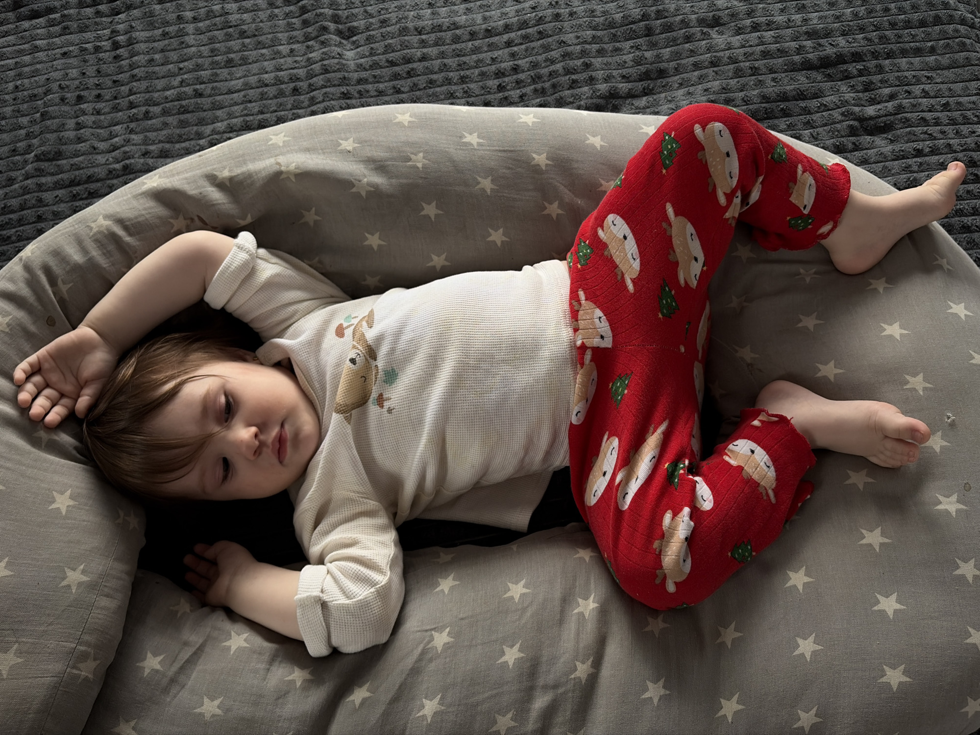 A toddler is lying on a grey nursing pillow with white stars. The pillow is placed on a grey textured blanket. The child is wearing a white long-sleeved shirt with a deer print and red Christmas-themed pants. The pants have reindeer and Christmas tree designs. The child is lying on their side with one arm outstretched and the other bent. The setting is indoors. The location is unknown and cannot be assumed to be in Chaikovsky, Russia.