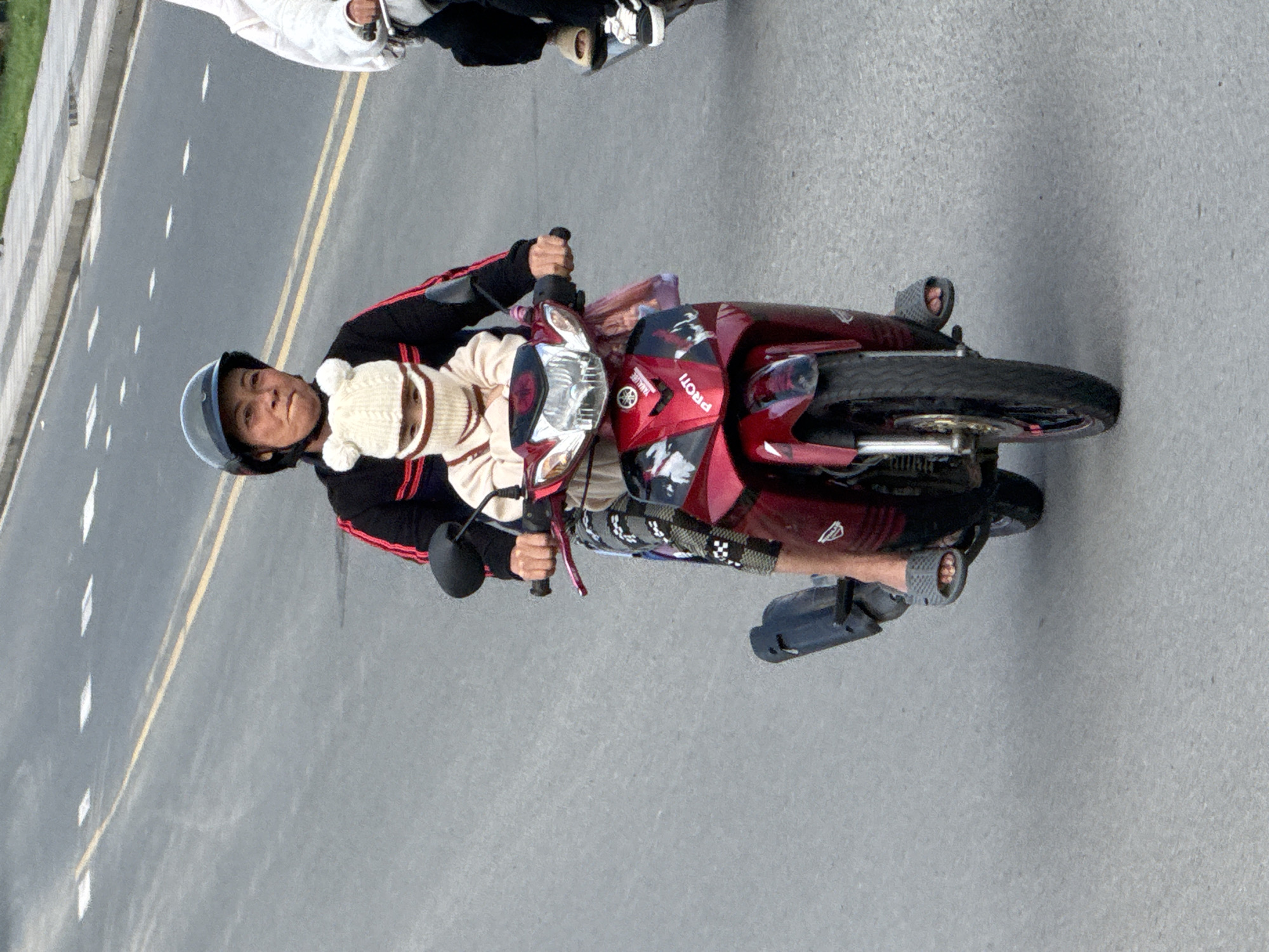 The image shows a woman riding a red motorcycle on a paved road in Da Lat, Vietnam. She is wearing a black jacket with red stripes and a black helmet. A young child, dressed in a beige sweater and a white knitted hat with ear coverings, is seated in front of her. The child's face is covered by the hat and a striped face mask, with only their eyes visible. The motorcycle has a prominent "PROTI" logo on the front. The road appears to be a typical urban street with visible lane markings. Another motorcycle with a person on it is partially visible to the right of the frame. The scene captures a common mode of transportation in Vietnam.