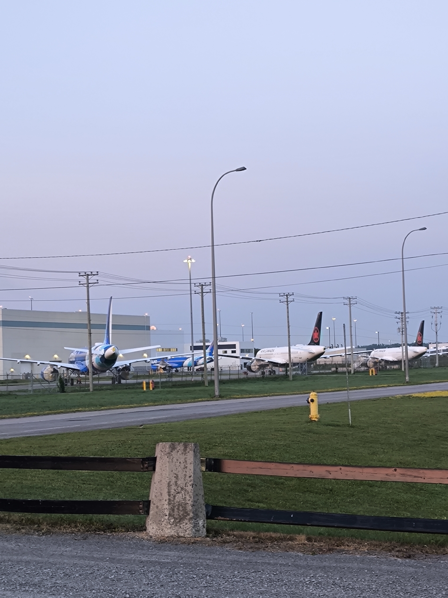 The image depicts an outdoor scene at the Montreal–Mirabel International Airport in Mirabel, Canada. The sky is a pale, dusky blue, suggesting either dawn or dusk. In the mid-ground, several commercial aircraft are parked on the tarmac. Two aircraft with blue and white livery are on the left, with one prominently displayed from the rear, showing its tailfin. To the right, two white Air Canada aircraft are parked, their red maple leaf logos visible on the tailfins. A large, light-colored building, possibly a hangar or terminal, is situated behind the aircraft on the left. Overhead power lines crisscross the scene, supported by wooden utility poles. Streetlights are also visible, some illuminated. In the foreground, a wooden fence with two dark horizontal planks is in sharp focus. Beyond the fence is a grassy area, and further back, a paved road or taxiway. A bright yellow fire hydrant stands on the grass in the middle right of the frame. The ground in the immediate foreground is covered in grey gravel. There are no discernible people or animals in the image.