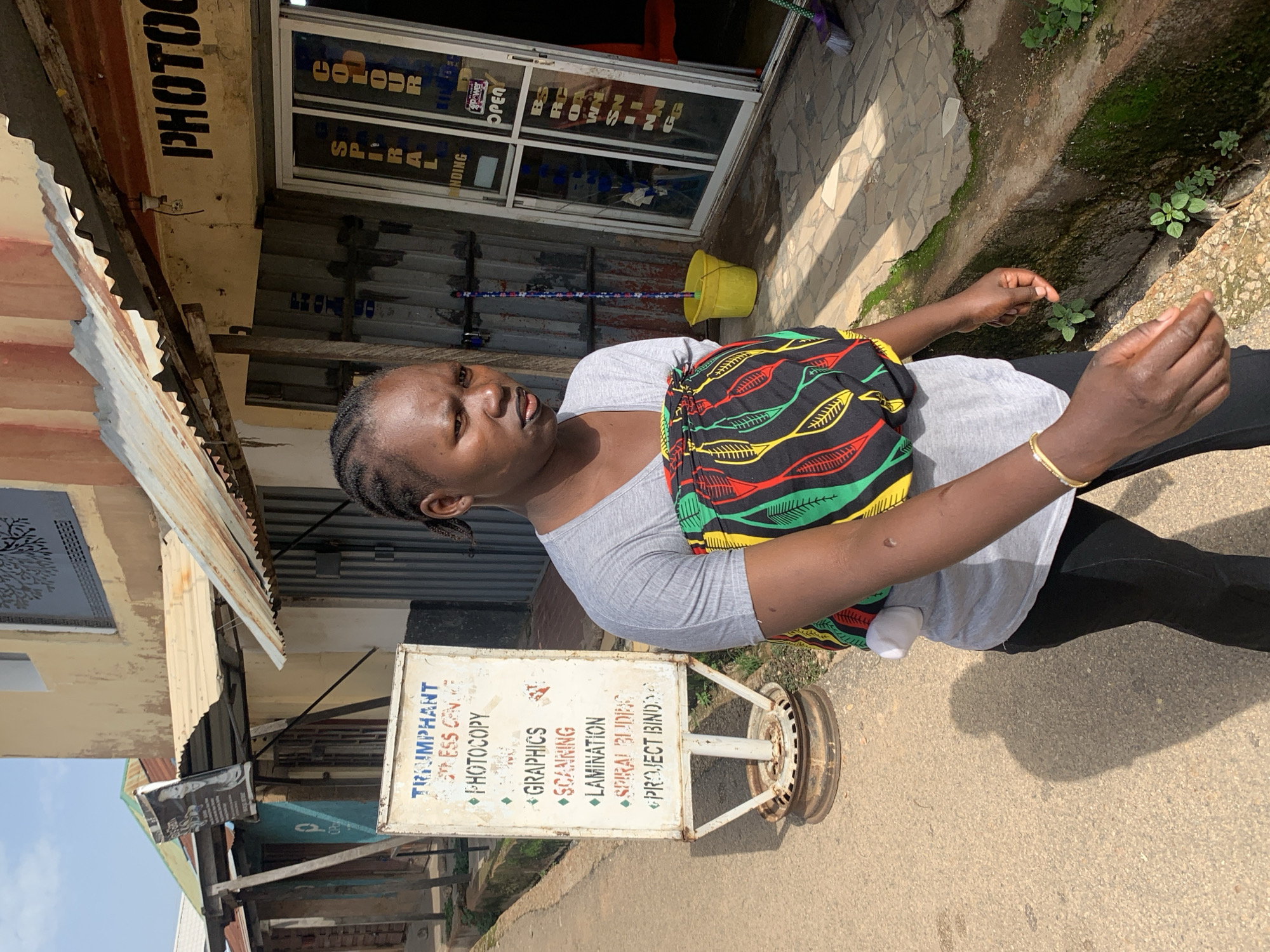 A dark-skinned woman with braided hair and dark lipstick walks past a shop on a sunny day. She is wearing a grey t-shirt and black pants, with a colorful fabric wrapped around her waist like a baby carrier. The shop's facade features a corrugated iron roof and windows displaying signs for "PHOTOCOPY," "SPIRAL BINDING," and "COLOUR PRINTING." A yellow bucket and a mop stand outside the shop. A white signpost with a list of services, including "PHOTCOPY," "GRAPHICS," "SCANNING," "LAMINATION," "SPIRAL BINDING," and "PROJECT BINDING," is also visible. The ground is paved with stone tiles in front of the shop and asphalt further down the street. The overall scene suggests a commercial area in Jos, Nigeria, with everyday business activities taking place.