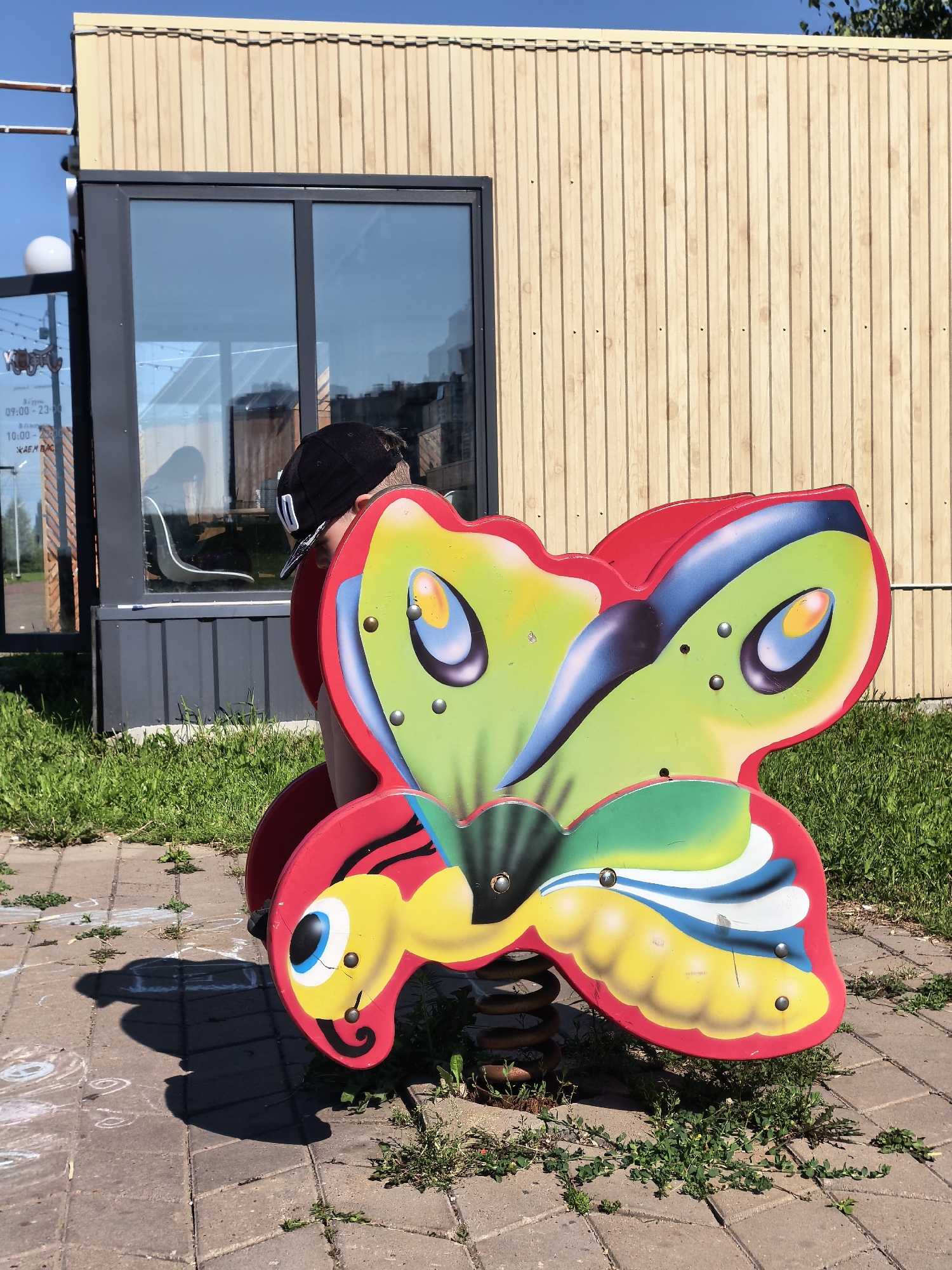 A young person wearing a black baseball cap is partially obscured behind a brightly colored, spring-mounted playground toy shaped like a butterfly. The toy is red, yellow, green, and blue, with large, cartoonish eyes. The scene appears to be an outdoor urban environment, likely a playground or public space, with a modern building with vertical wood paneling and large windows in the background. The ground is paved with interlocking bricks and scattered with patches of green grass and weeds. Sunlight casts a shadow of the butterfly toy on the ground, indicating it is a sunny day. The building in the background has reflective windows that show glimpses of the interior and the surrounding environment. The presence of a building with signs of business hours (visible on the window) suggests a commercial or public establishment in Sterlitamak, Russia.