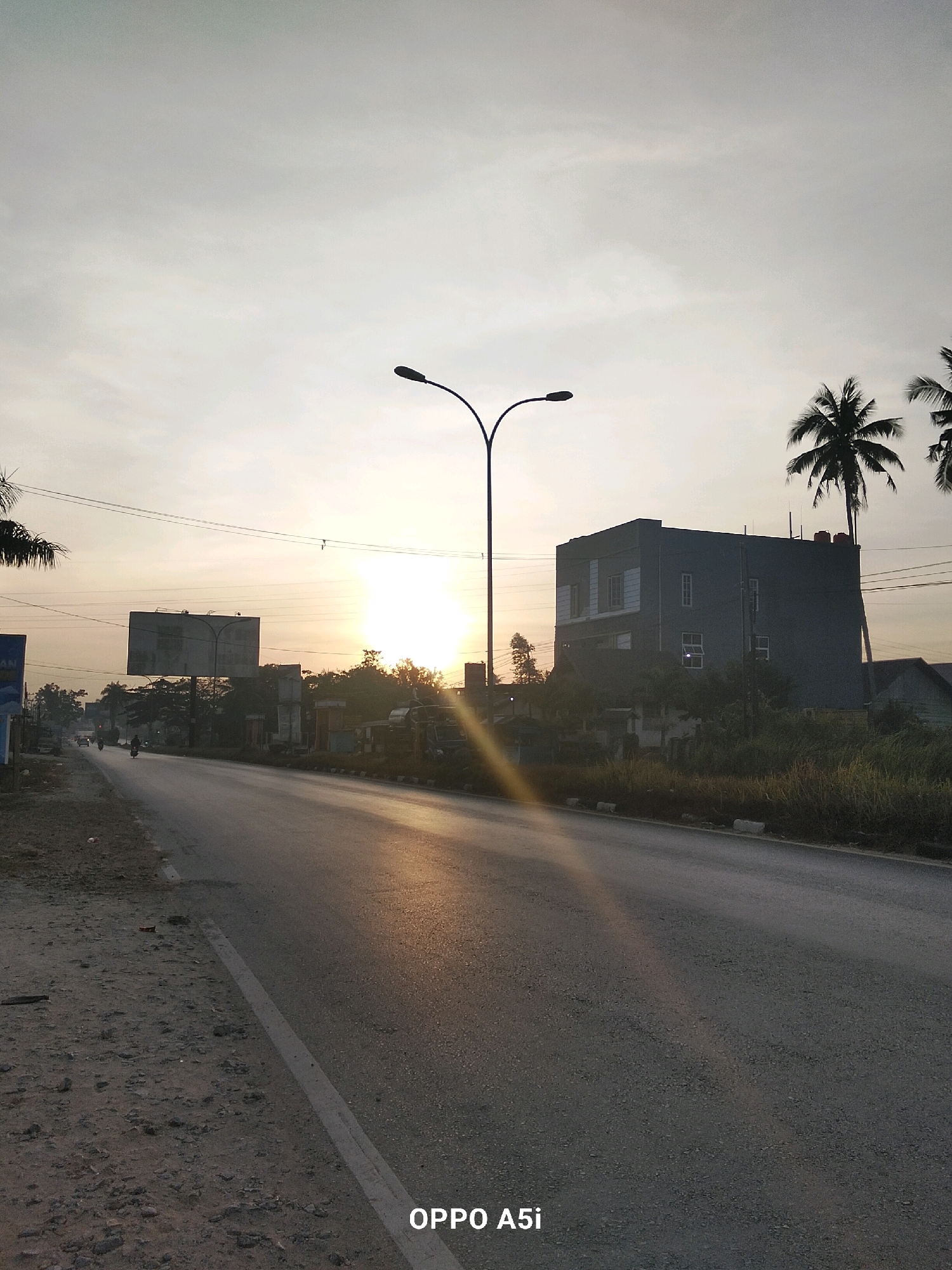 The image depicts a street scene in Watubangga, Indonesia, at sunrise. The sun is low on the horizon, casting long shadows and a warm glow across the asphalt road. Two streetlights with curved poles are prominent in the foreground. On the right side of the road, a modern, multi-story building with a dark facade and several windows stands out, with palm trees in front of it. On the left side of the road, a billboard is visible, along with a few motorcycles and what appear to be other buildings in the distance. The sky is hazy with a pale yellow and grey hue. The road itself is paved, with a white line marking the edge of the lane. The area to the left of the road appears to be a dirt or gravel shoulder. A watermark at the bottom right corner reads "OPPO A5i".
