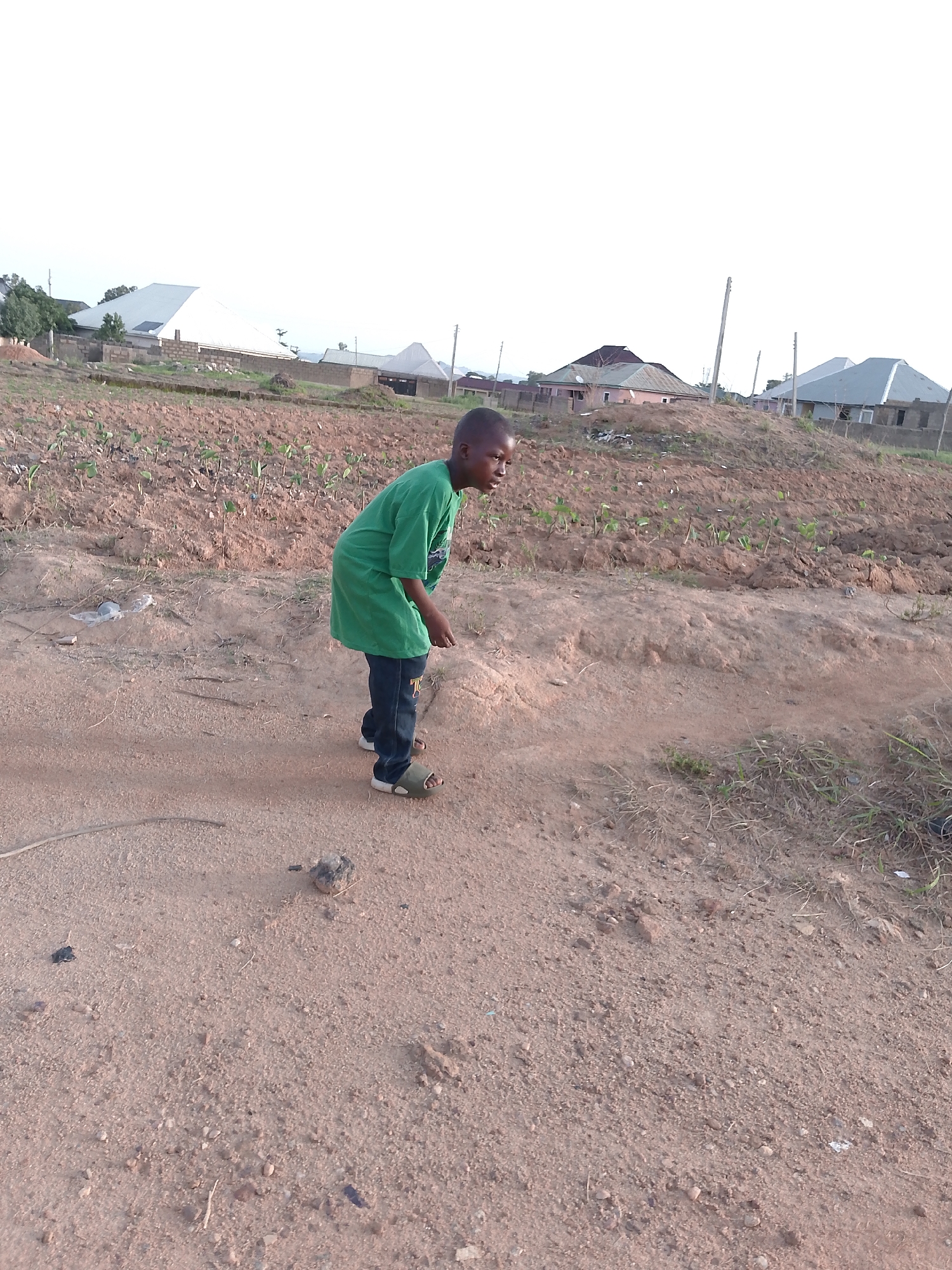 A young boy wearing a green t-shirt and blue jeans with sandals is standing on a dirt path in what appears to be a rural or semi-rural area. Behind him is a field with rows of young plants, likely crops. In the background, there are several houses with corrugated metal roofs, suggesting a residential area. The sky is overcast, and the overall lighting suggests it is daytime. The environment consists of dry, reddish-brown soil and sparse vegetation. The image appears to be taken in Barikin Gidar Kanat, Nigeria.