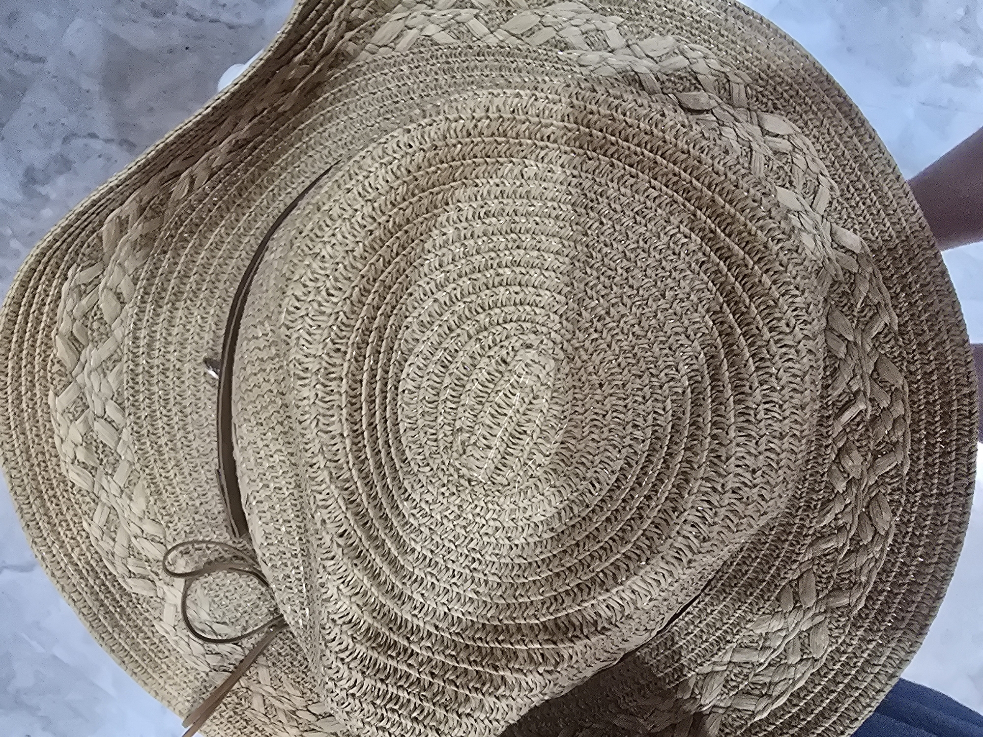 A close-up shot focuses on a woven straw hat, likely for sun protection. The hat is a light brown color and features a braided band around the brim, with a thin brown ribbon tied in a bow on the crown. The texture of the straw is clearly visible, showing the intricate weaving pattern. The hat is resting on an unseen surface, with a hint of a pale blue and gray background that appears to be a wall or textured ground. The lighting suggests it is daytime. There are no discernible landmarks or elements that would place this image specifically in Ulan-Ude, Russia, or any other city. The image appears to be a still life focusing solely on the hat.