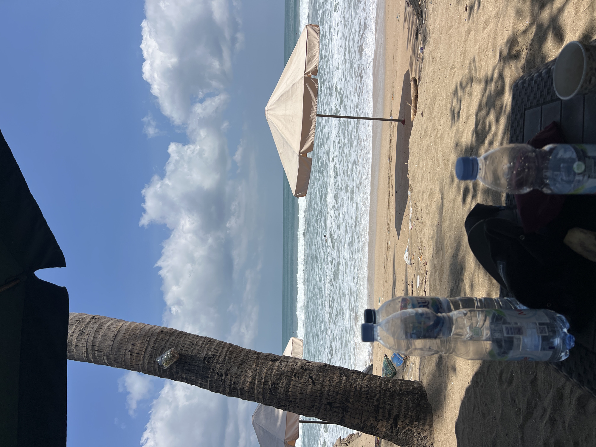 The image depicts a beach scene in Kuta, Indonesia, on a sunny day with scattered clouds. In the foreground, a textured surface, likely a table, is visible, with two plastic water bottles and a dark bag or fabric resting on it. Another water bottle is positioned further back. A palm tree trunk stands prominently on the left side of the frame, with a plastic bottle attached to it. The sand beach stretches towards the ocean, where moderate waves are breaking. Two beige beach umbrellas are set up on the sand, one closer to the foreground and another further back. In the distance, a small figure is in the water, possibly swimming or surfing. The overall environment is a typical beach setting, suggesting a leisure or vacation context.