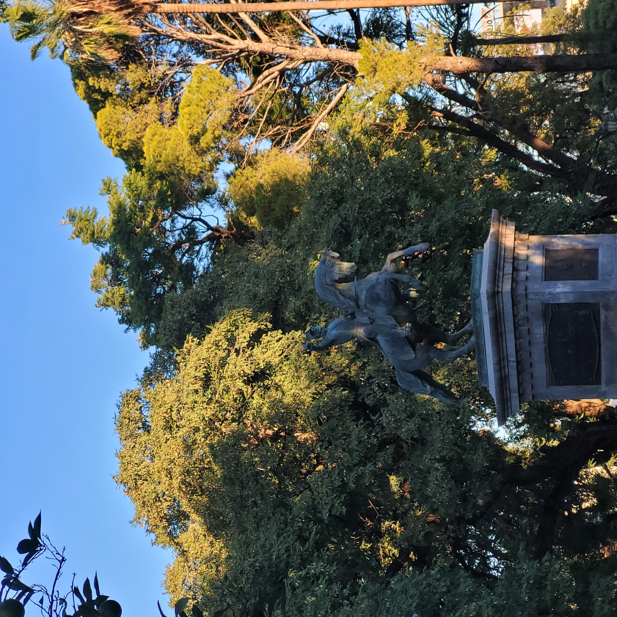 The image shows a bronze equestrian statue on a stone base, set among trees in a park. The statue depicts a rider on horseback, with the horse rearing up. The base has rectangular panels on its sides. The sky is visible in the upper left corner, and parts of buildings are faintly visible through the trees. This statue is located in San Fernando del Valle de Catamarca, Argentina.