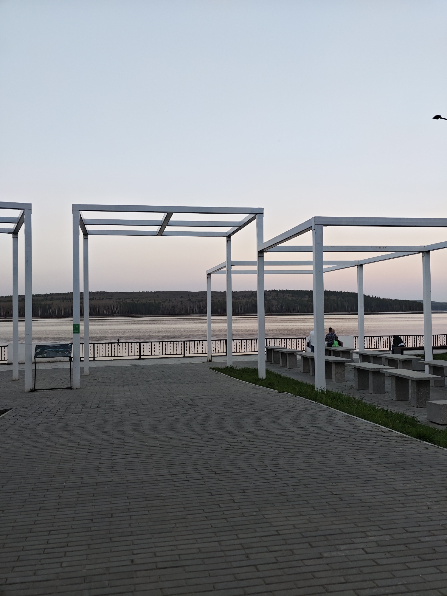 This image shows a waterfront park or promenade in Chaikovsky, Russia, at dusk. Four white, cubic metal frames stand along a gray brick walkway. A low, black metal fence separates the walkway from the water, with trees visible on the opposite shore. Several concrete picnic tables with benches are positioned near the metal frames. Two people are sitting at a table under one of the frames. A trash can is beside the table. A sign is next to one of the frames. A lamp post is visible in the sky at the upper right of the image.