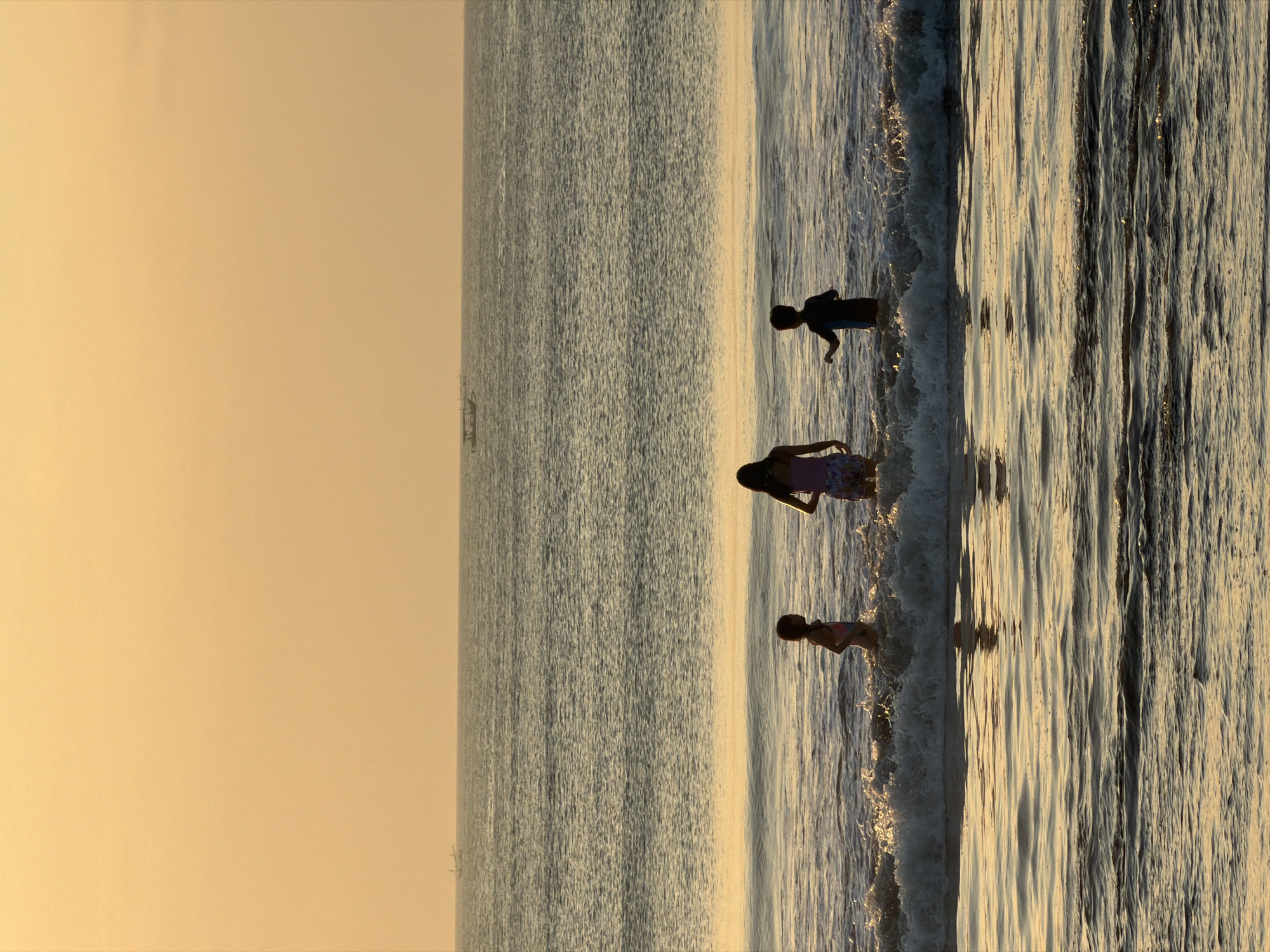 A vertical, full-frame, medium-shot photograph captures three children wading in the ocean waves at sunset on Kuta Selatan beach, Bali, Indonesia. The sky is a hazy gradient of soft orange and yellow, with two small fishing boats visible on the horizon in the distance. The sea is a textured expanse of gray-blue water, reflecting the warm hues of the sky. The children are silhouetted against the water and sky, with their forms dimly discernible. The child on the left, appearing to be a girl, wears a swimsuit and stands facing left. The child in the center, also a girl, wears a patterned dress and faces forward, her arms slightly outstretched. The child on the right, a boy, wears a dark, short-sleeved shirt and shorts and faces towards the center child, with one arm raised. They are all standing amidst breaking waves that create white foam and spray. The foreground shows calmer water with the reflection of the sky and the silhouettes of the children distorted by the gentle ripples. The overall mood of the image is serene and tranquil, evoking a sense of childhood play at the end of the day.