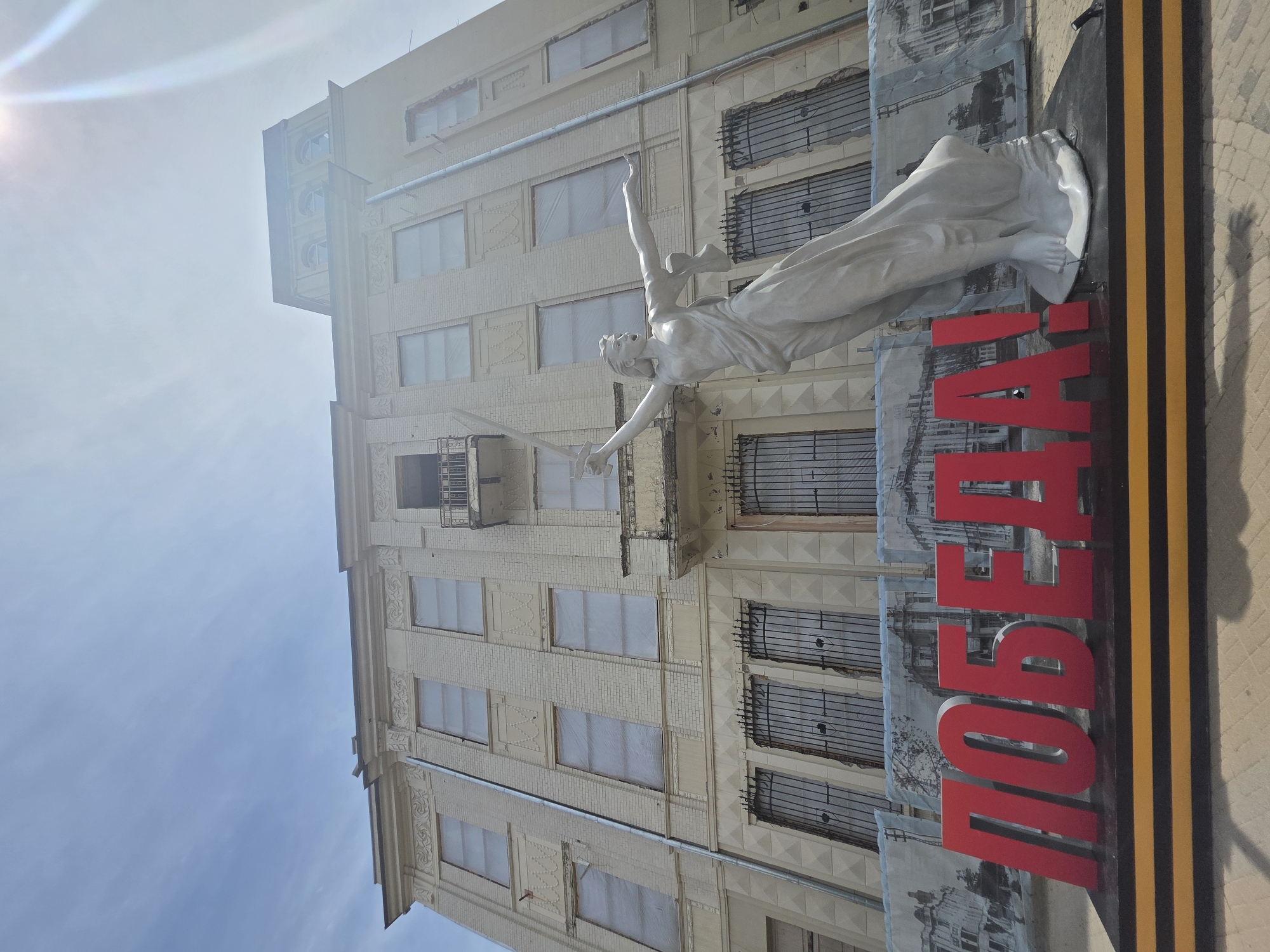 The image captures a monument in Krasnodar, Russia. A white statue of a female figure, possibly winged, with a sword in hand, is prominently displayed alongside a sign with red letters that spell out "ПОБЕДА", which translates to "Victory". The monument appears to be placed against the backdrop of a multi-story, light-colored building with several windows. Some windows seem to be covered. The building's façade shows architectural details. The overall setting suggests an urban environment during daylight, with clear skies above.
