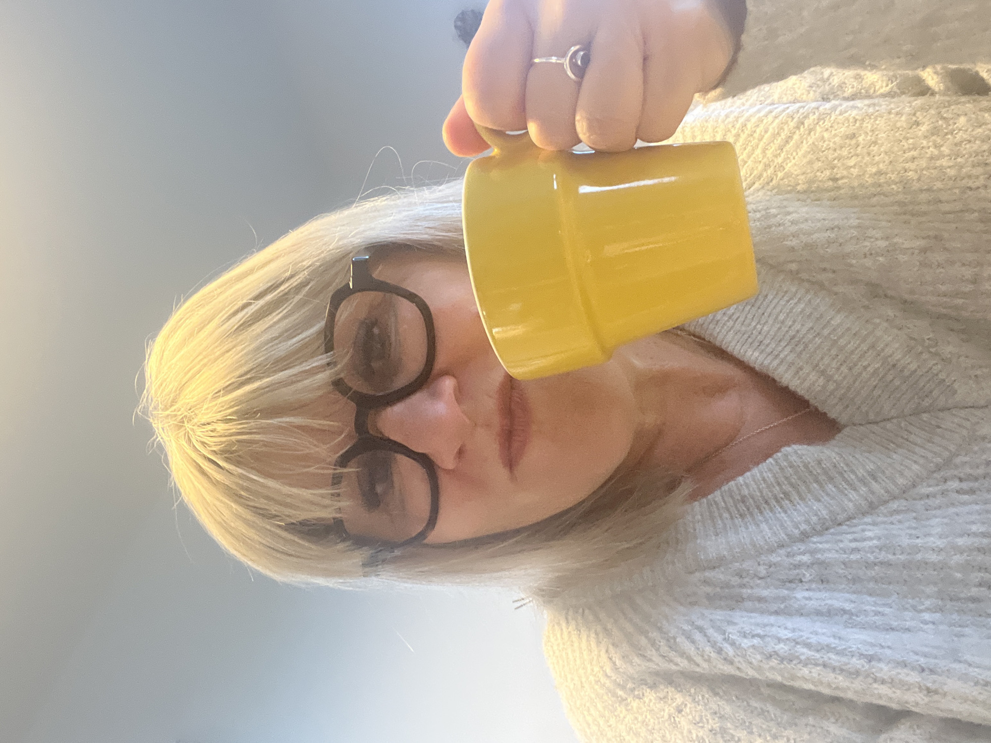 The image, captured in Chambœuf, France, depicts an adult individual in a close-up, frontal composition. The subject has blonde hair with bangs, wears dark, round-framed eyeglasses, and is dressed in a light-colored, V-neck chunky knit sweater. A thin silver chain necklace is visible. The subject's right hand holds a bright yellow, ridged mug to the mouth, indicating an action of drinking or preparation to drink, with lips slightly pursed. A silver ring featuring a dark stone is present on the right ring finger. The background is an indistinct, uniformly light grey or bluish-grey field, lacking specific environmental identifiers.