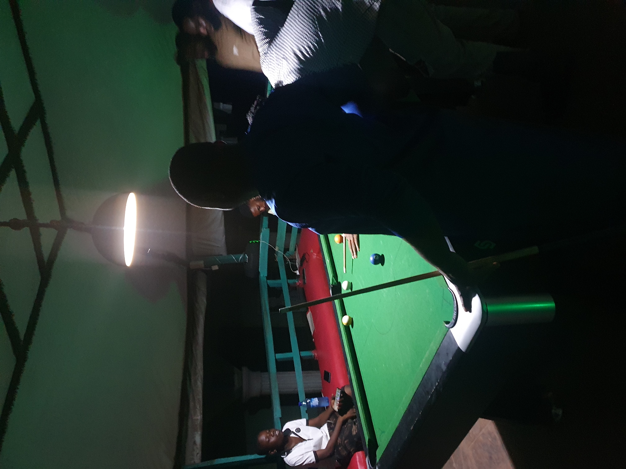 This image depicts a dimly lit indoor scene, likely a recreational area, with a pool table as the central focus. The environment is characterized by a green-colored pool table with several billiard balls scattered on its surface, including a blue cue ball. A pool cue rests on the table, held by an unseen individual.
In the foreground, a person's back is visible, silhouetted against the ambient light, suggesting they are playing pool. To the left, a young Black male is seated on a red bench beside the pool table, looking away from the camera. He is wearing a white polo shirt with a dark collar and camouflage-patterned shorts, and he is holding a mobile phone. A clear plastic bottle of water is also present on the bench.
In the background, several other individuals are partially visible, appearing to be standing or gathered around the pool table, some with their faces obscured by shadows. The ceiling above is dark and appears to be a canopy structure with exposed beams. A prominent light fixture hangs from the ceiling, casting a bright light that illuminates the pool table and parts of the surrounding area. The overall atmosphere suggests a casual gathering or game night. The specific location in Lokoja, Nigeria, cannot be determined from the visual information provided in the image.