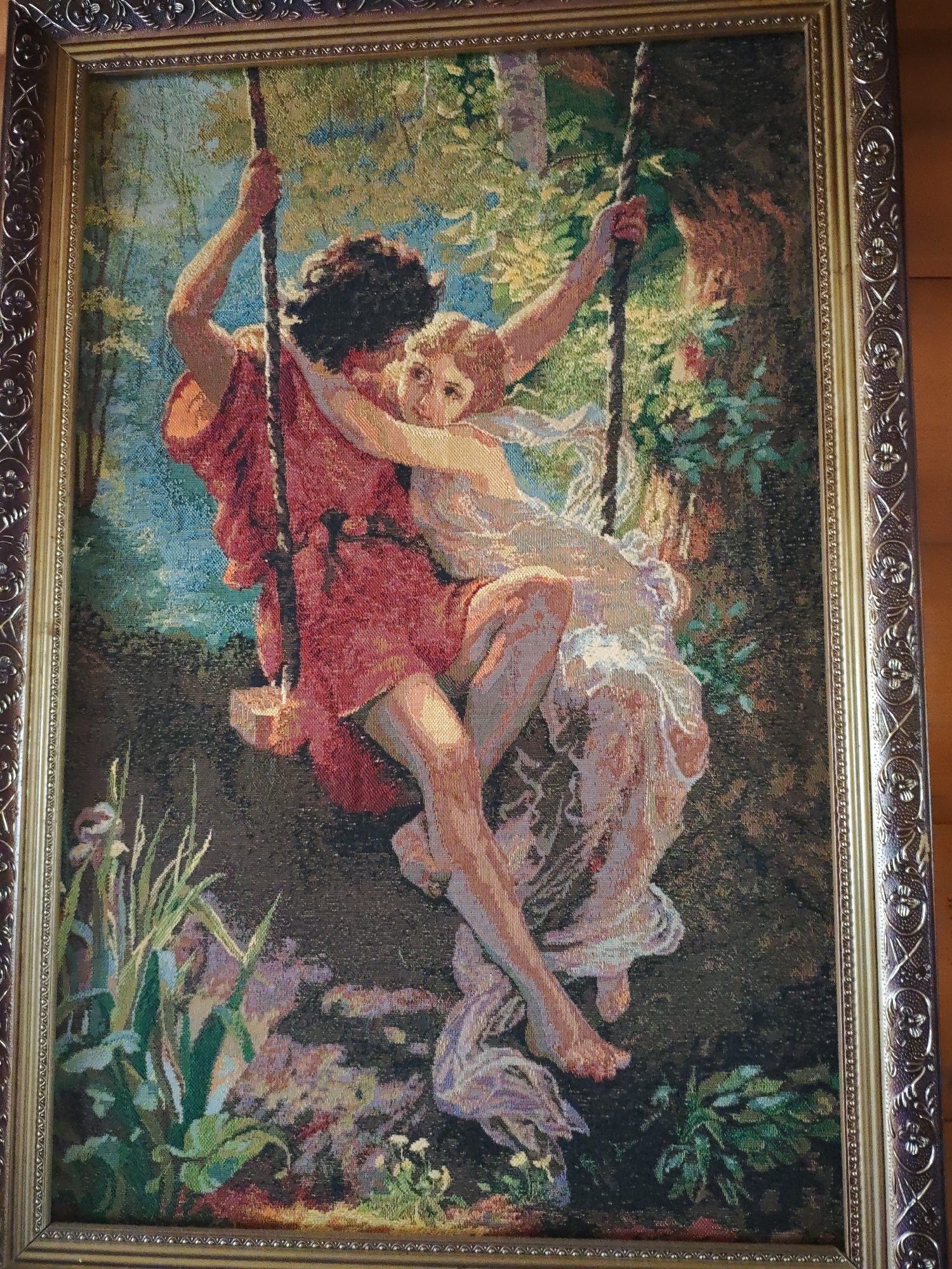 A framed tapestry depicts a man and a woman on a swing in a forested area. The man, with dark hair, is wearing a red garment and holds the swing ropes. The woman, with light hair, is clad in a white dress and is embracing the man. The background features trees, foliage, and a glimpse of blue sky. In the foreground, there is greenery, including what appear to be irises. The location is unknown, and there's no evidence to connect the image to Боровково, Russia.