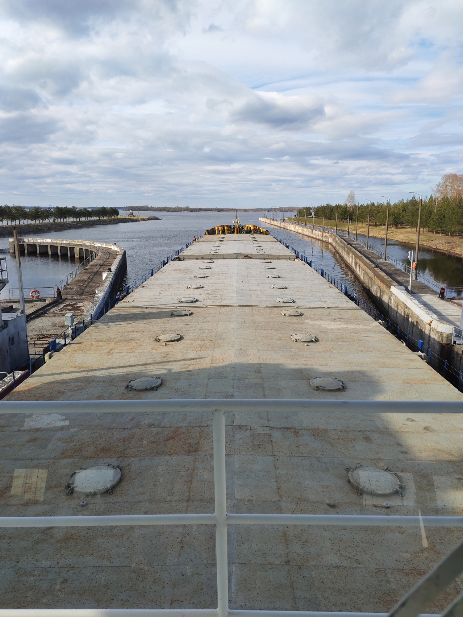 A high-angle, outdoor photograph shows the deck of a large vessel passing through a canal lock in Unknown, Unknown. The vessel's deck, a textured gray surface with numerous circular access hatches, dominates the foreground, partially obscured by a white railing. Beyond the deck, the canal stretches into the distance, flanked by concrete walls on both sides. On the right, a person is visible standing by a bollard, while several other people appear indistinctly towards the end of the vessel. The canal walls are lined with evenly spaced trees. The sky is overcast with predominantly gray clouds. Water fills the canal and the body of water it connects to, visible in the background.