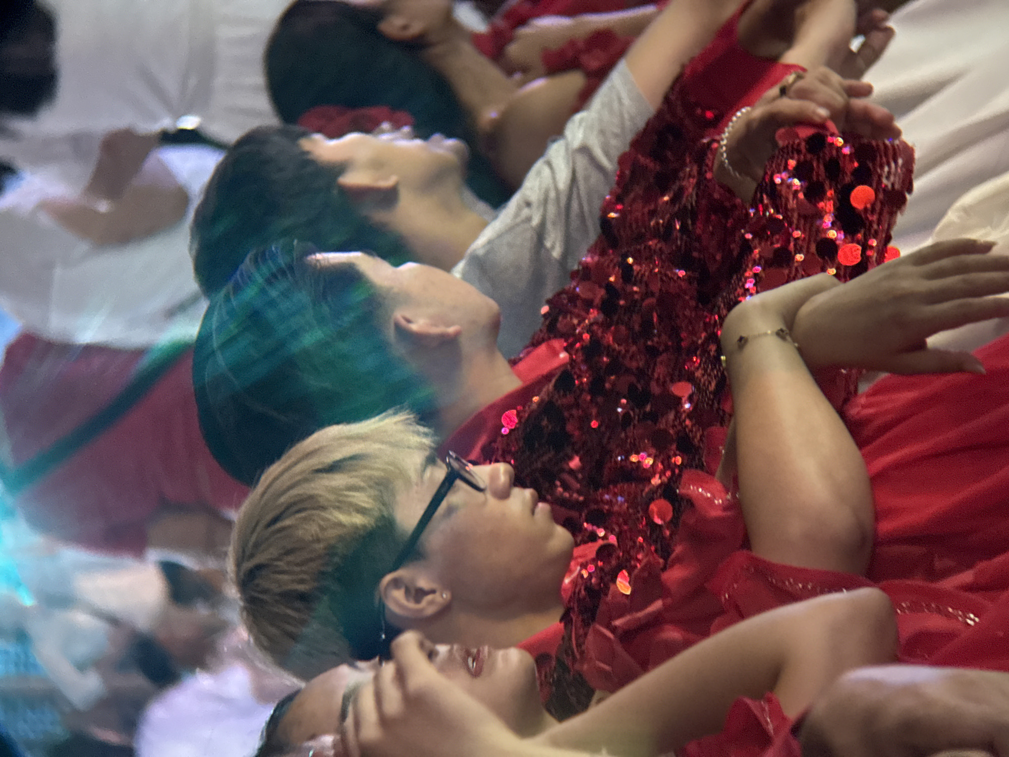 The image captures a group of people in what appears to be a performance or festival setting. The foreground features a person wearing a red, sequined garment. Their left arm is visible, adorned with a bracelet. To their right, another individual is seen from the side, wearing glasses and also in red attire. Further in the background, other individuals are present, some in white clothing, and there are hints of bright lighting. The scene suggests a lively, possibly outdoor, event. This photograph was taken in Nha Trang, Vietnam, likely during a cultural celebration or performance.