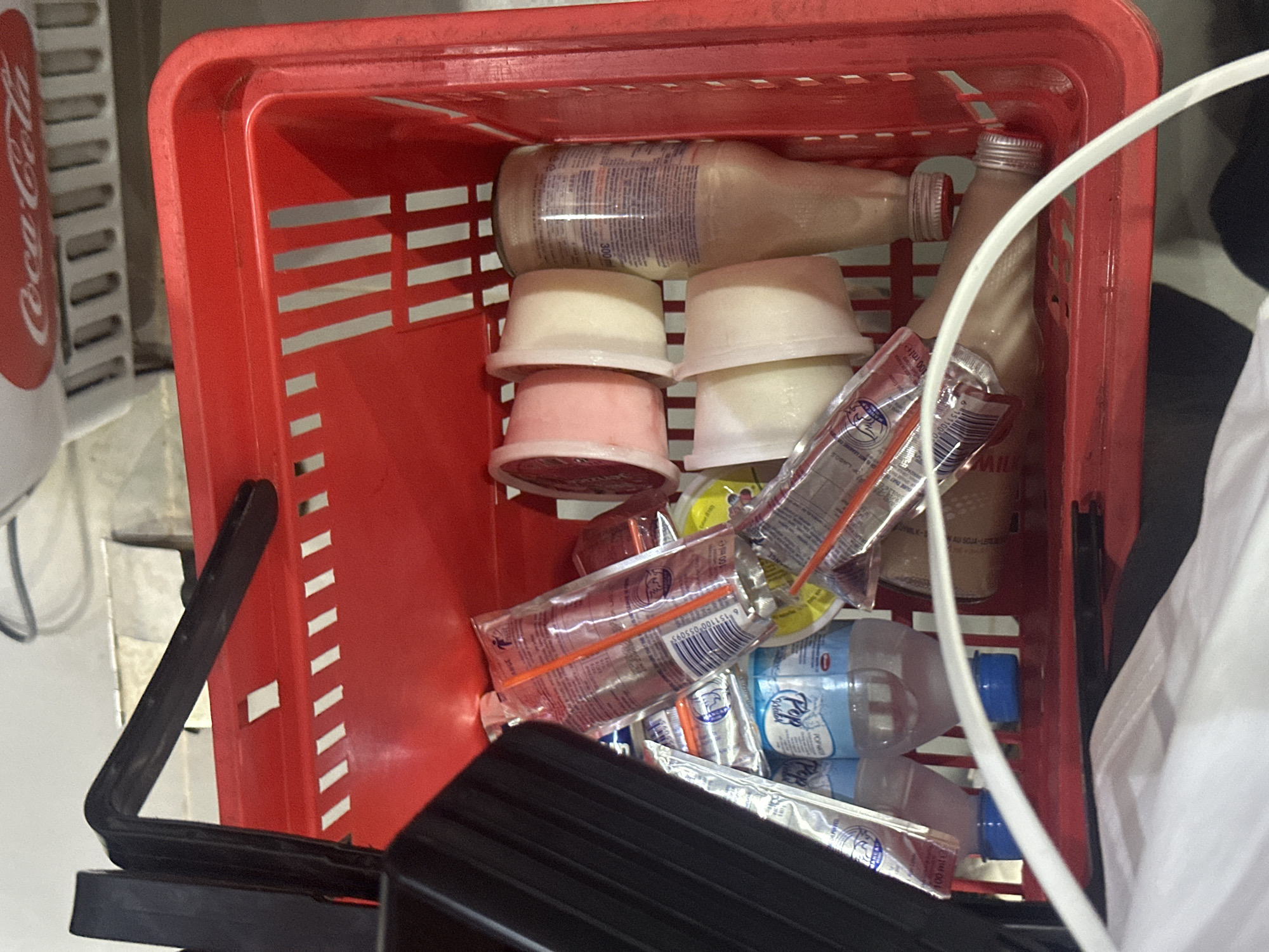 A red plastic shopping basket is filled with assorted food and beverage items. These include two glass bottles of milk, several small plastic tubs containing what appears to be yogurt, a package of juice boxes with straws, and some small water bottles. The shopping basket sits in a shopping area, with a cooler for Coca-Cola visible nearby. A portion of a white shirt can be seen in the right foreground of the image.