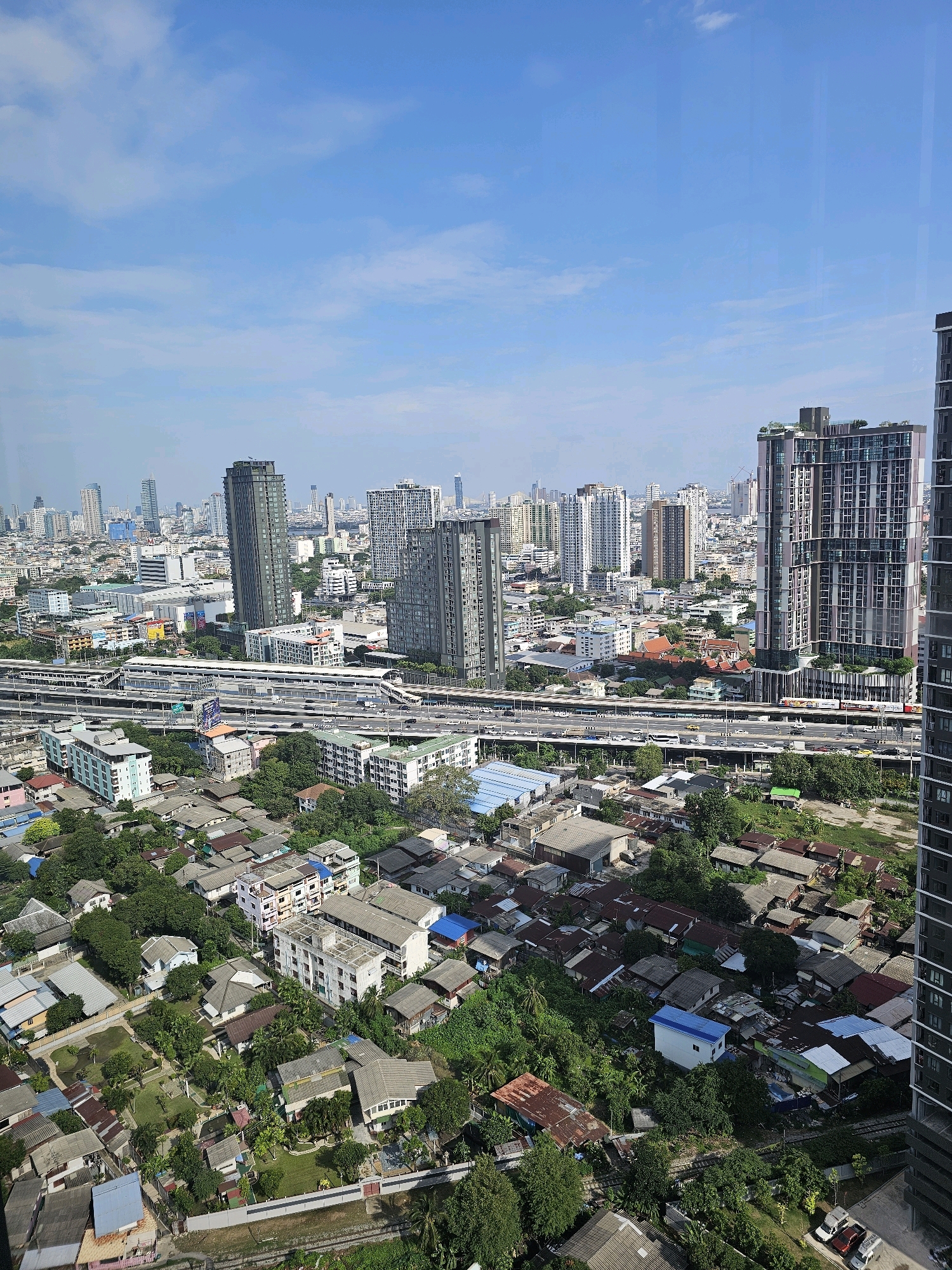 This is a high-angle, medium-shot, eye-level, vertical photograph of a dense urban landscape in Bangkok, Thailand, under a partly cloudy blue sky. The foreground features a lower-lying residential area with numerous houses, many with tin roofs, interspersed with lush green trees and vegetation. A network of railway tracks runs through this area. In the midground, a multi-level elevated highway system is visible, carrying traffic. Several modern, tall buildings, including skyscrapers, are prominent in the background, forming the city's skyline. One very tall, dark skyscraper is on the far right edge of the frame, partially obscuring the view. A long, low-rise building with a train station appears to be situated on one of the elevated highway levels. The scene depicts a mix of old and new urban development, with older residential neighborhoods existing alongside modern high-rise structures and infrastructure. There are no people or animals clearly visible in the image. The overall impression is of a bustling, sprawling metropolis.