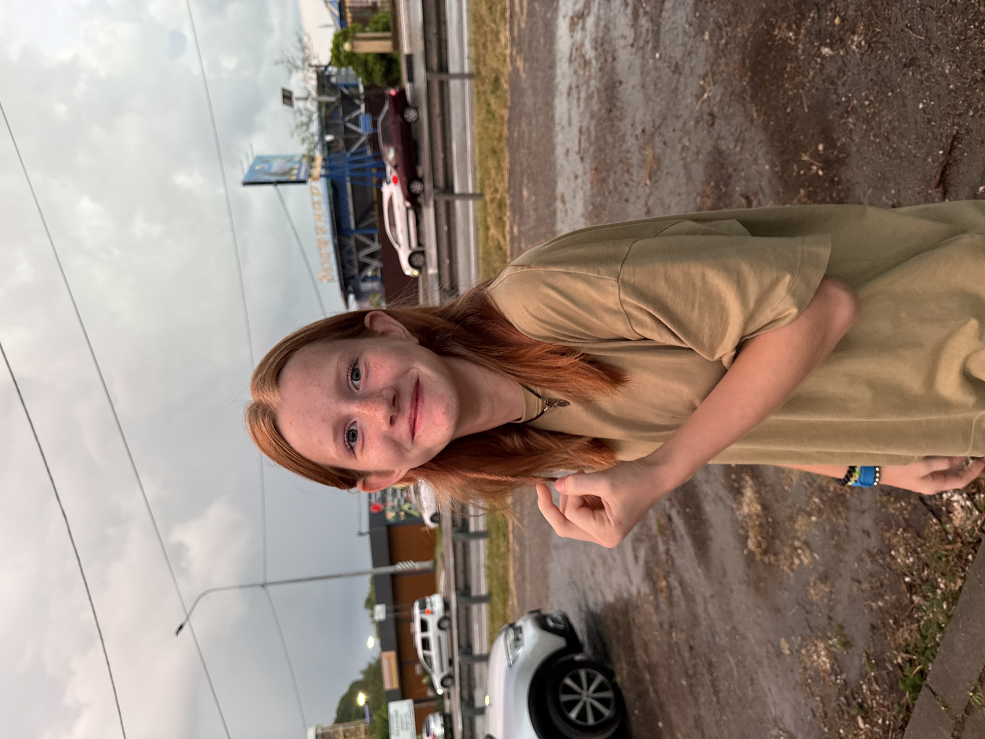 In the city of Nebug, Russia, a young Caucasian girl with red hair and freckles is captured in a medium shot, smiling at the camera. She is wearing a loose, tan t-shirt and a necklace with a pendant. Her right arm is bent, and her fist is clenched near her chest. The background reveals a street scene with cars, a guardrail, and a building with signage that is partially obscured. The ground appears wet, suggesting recent rain, and the sky is overcast with clouds. The overall atmosphere is subdued, possibly due to the weather conditions.