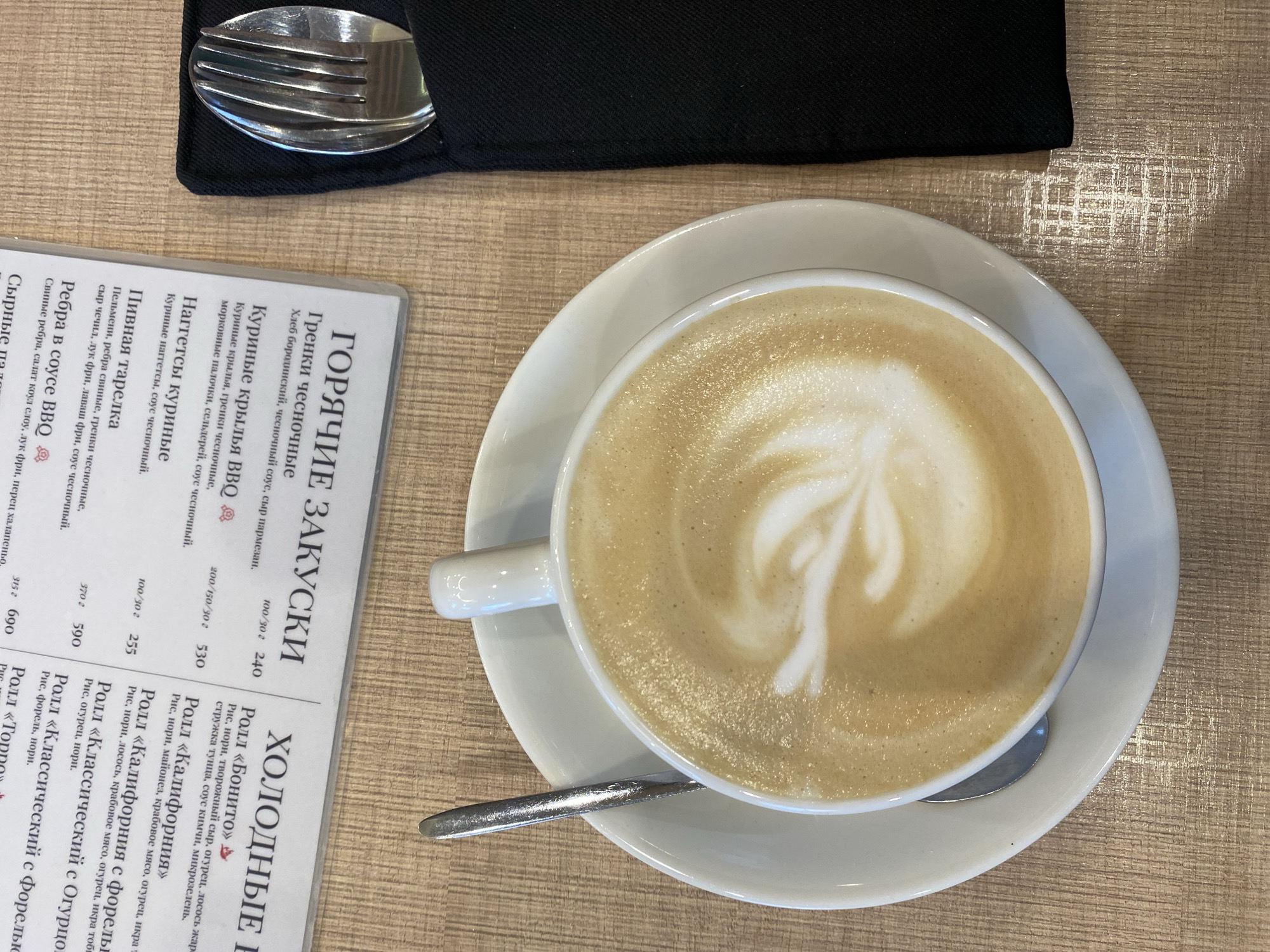 The image shows a top-down view of a table setting. On the table is a cup of coffee with latte art, a white saucer, a spoon, a black napkin with a fork, and a menu in Russian. The menu lists various appetizers, including hot and cold dishes such as chicken wings, nuggets, sushi rolls, and ribs. The table itself has a light brown, textured surface. No people are visible. The location information (Neftekamsk, Russia) is given but not evident from the image's content.