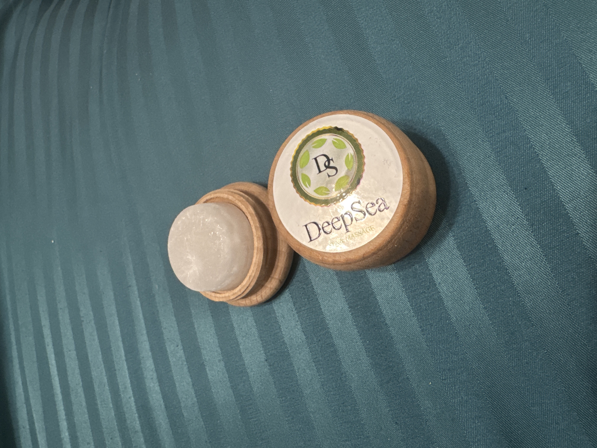 Two circular wooden containers rest on a teal-colored fabric surface. The fabric features a prominent striped pattern, with alternating glossy and matte bands running diagonally from the bottom left to the top right of the frame. One container is open, revealing a translucent, off-white, solid substance with a slightly textured, almost crystalline appearance, possibly a soap, balm, or scrub. The other container is closed and has a white circular label affixed to its top surface.
The label is decorated with a green border and small green leaf illustrations around its edge. In the center, there is a stylized dark green "DS" logo. Below the logo, the word "DeepSea" is printed in a dark, elegant script, followed by smaller text that reads "SPA & MASSAGE". The wooden containers are light-colored and appear to be made from a smooth, finished wood. The lighting is even, highlighting the textures of the fabric and the solid substance within the open container. No activity is taking place, and the setting appears to be an indoor environment.